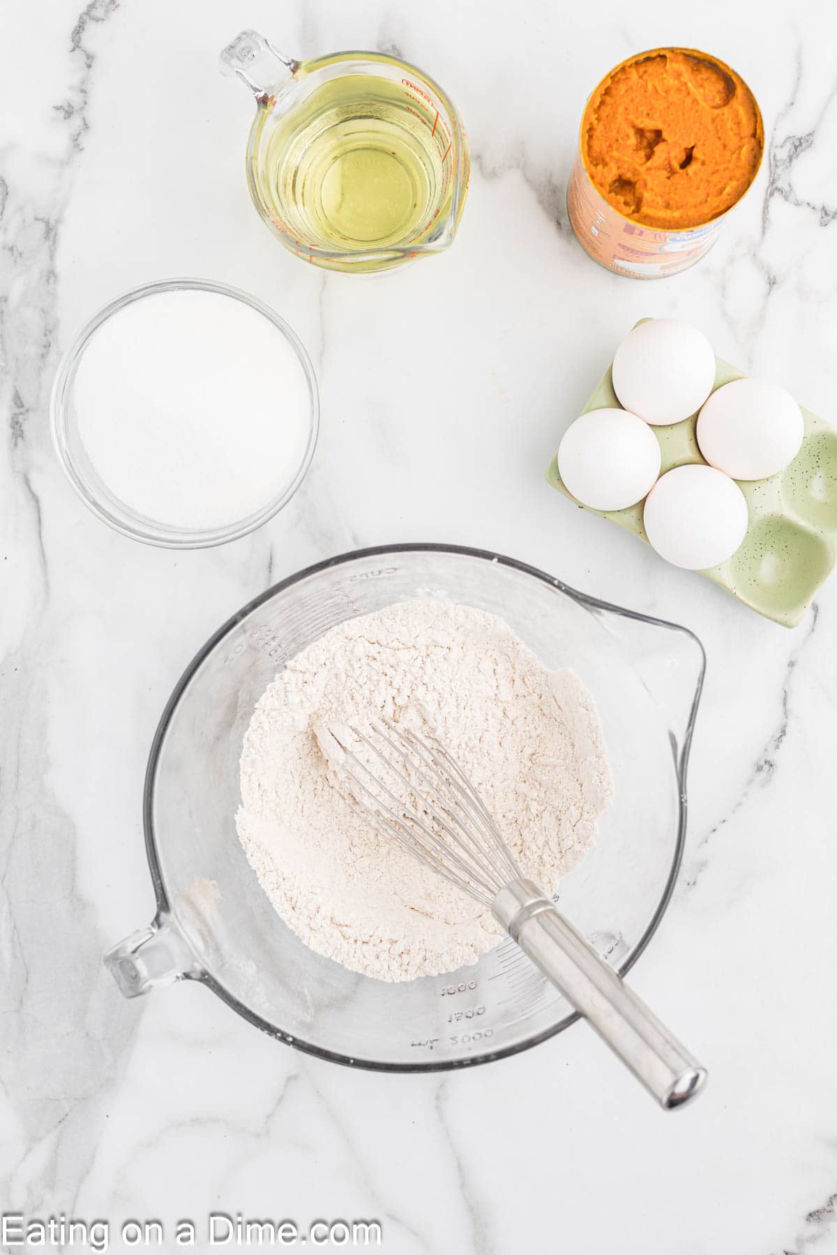 Whisking together dry ingredients in a bowl with a side of eggs in a carton, oil, and a can of pumpkin 