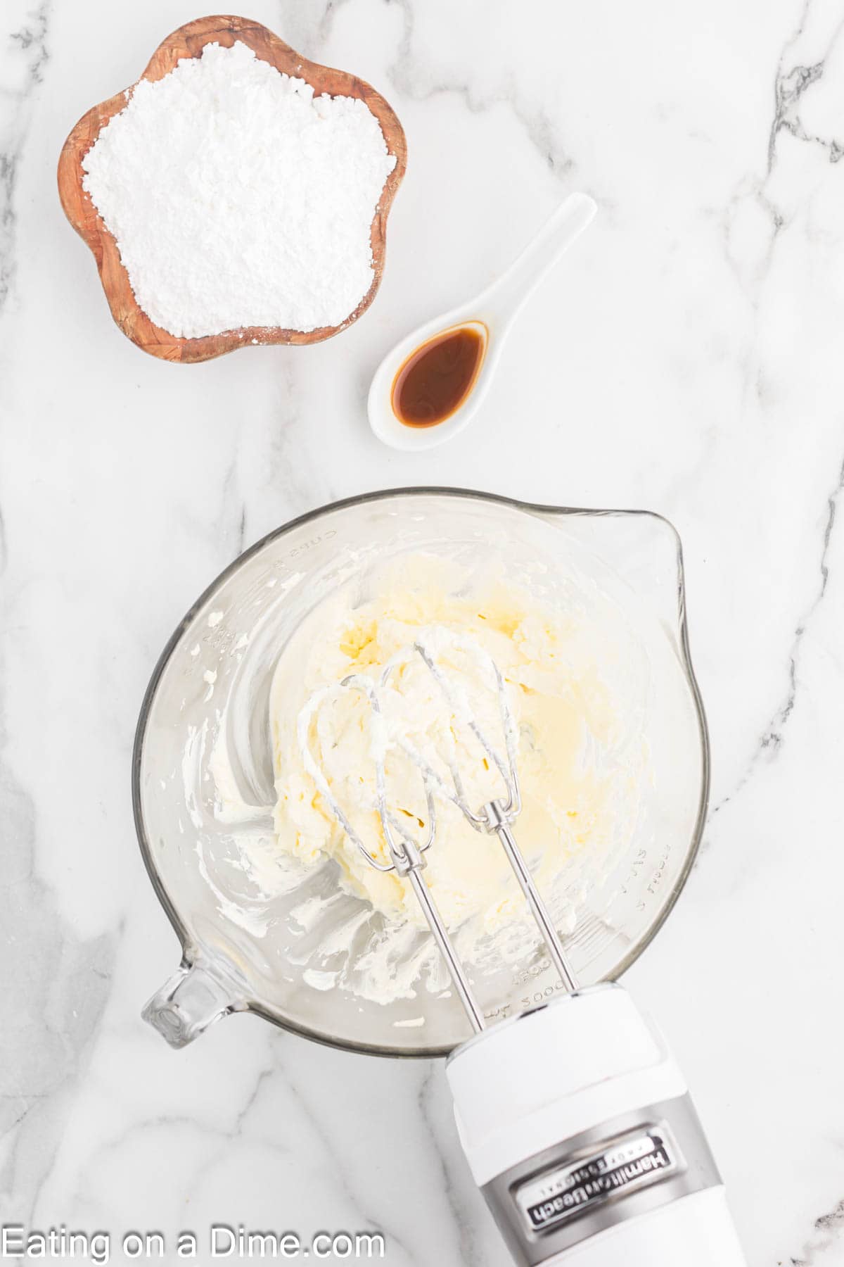 Beating cream cheese and butter in a bowl with a hand mixer with a side of vanilla extract in a teaspoon and a bowl of powdered sugar 