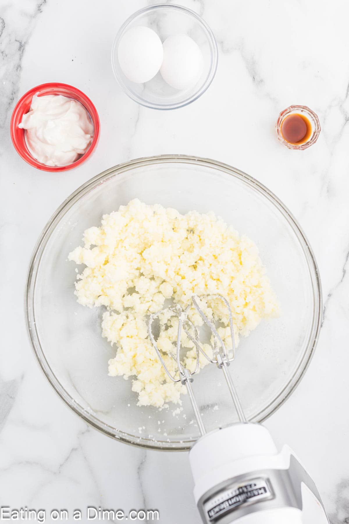 Beating sugar and butter together in a bowl with a hand mixer with small bowls of sour cream, eggs and vanilla on the side