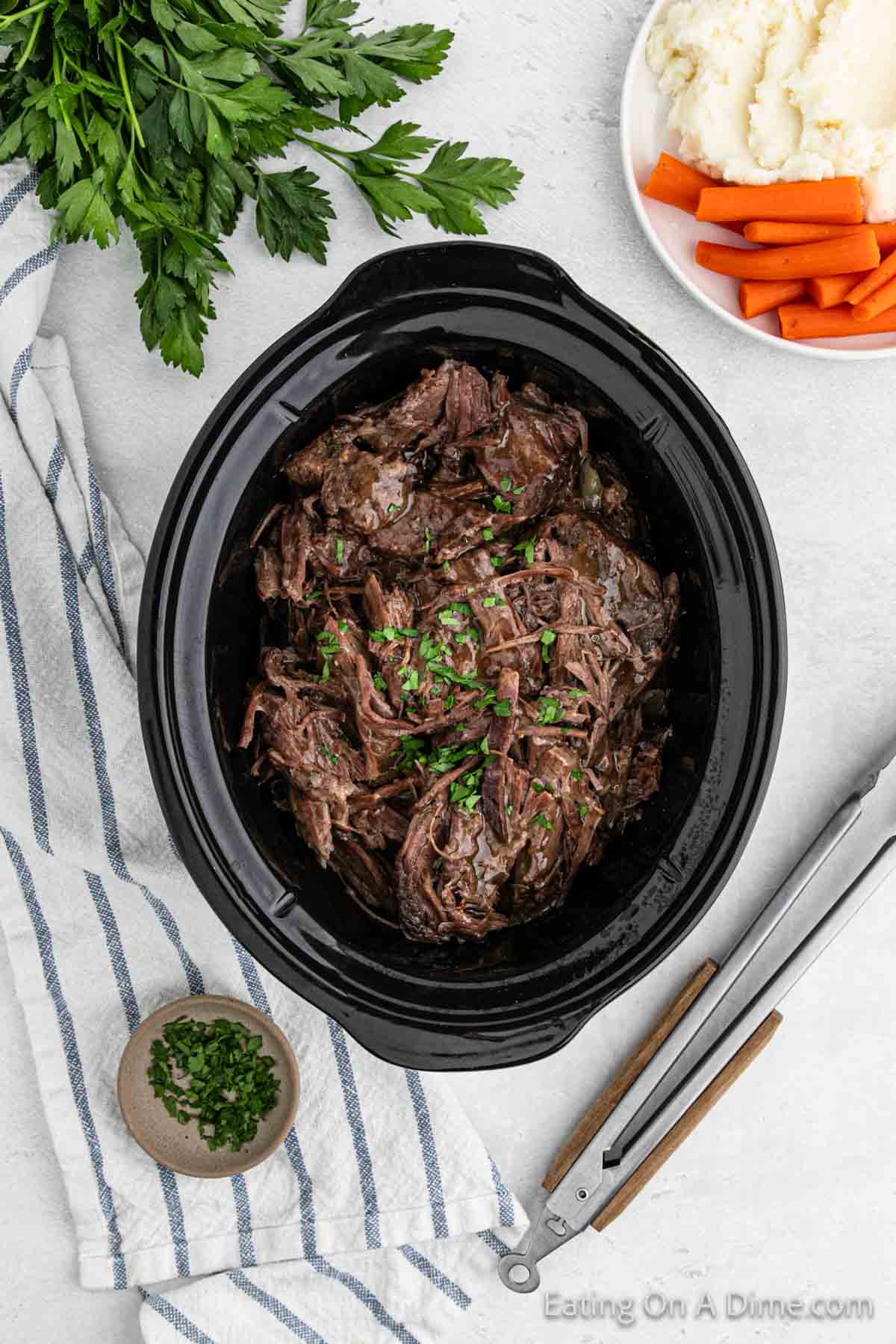 Shredded roast in the slow cooker with a plate of mashed potatoes and carrots on the side