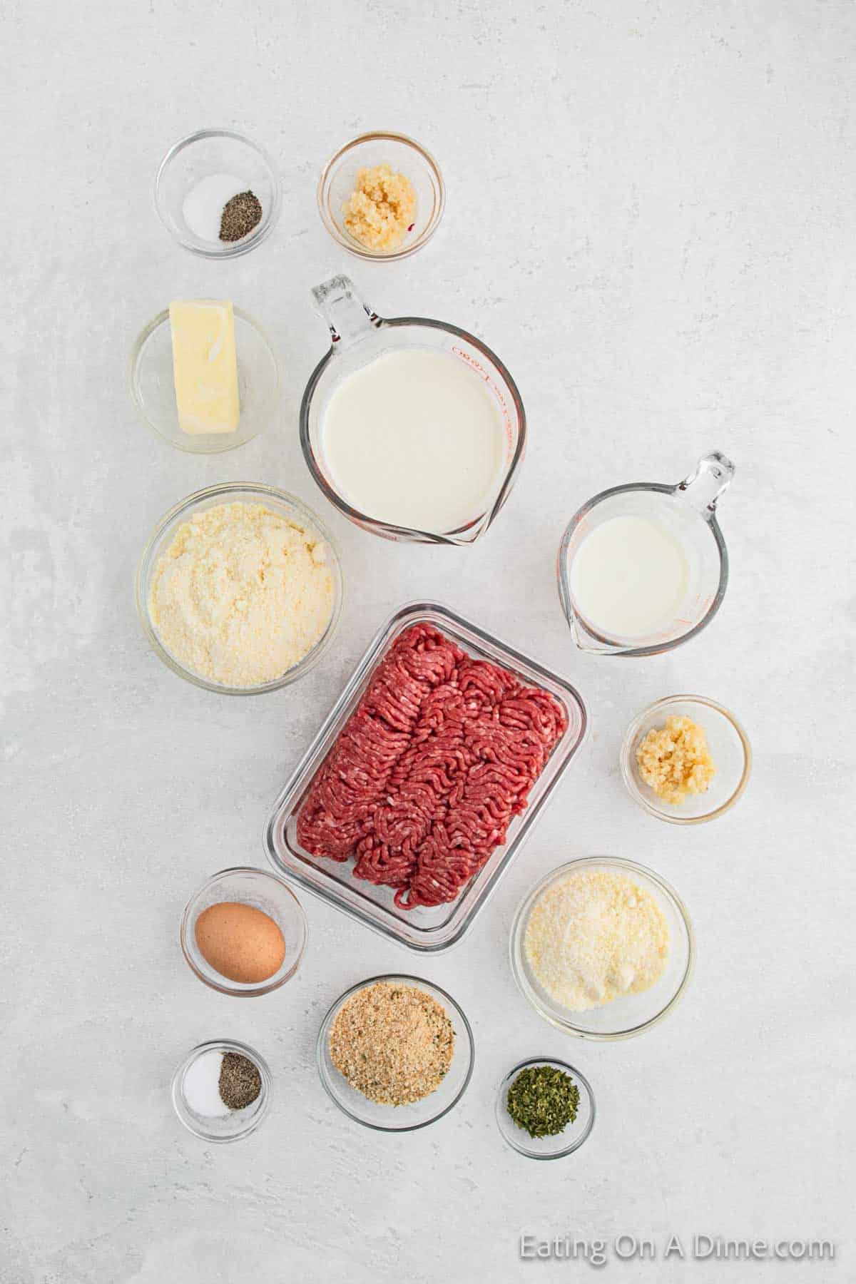 Ingredients for a recipe laid out on a light-colored surface. Includes ground meat for meatballs, grated cheese, dry breadcrumbs, seasonings, butter, milk, an egg, minced garlic, and other small amounts of spices and seasonings in bowls. Text reads "Eating On A Dime.com".