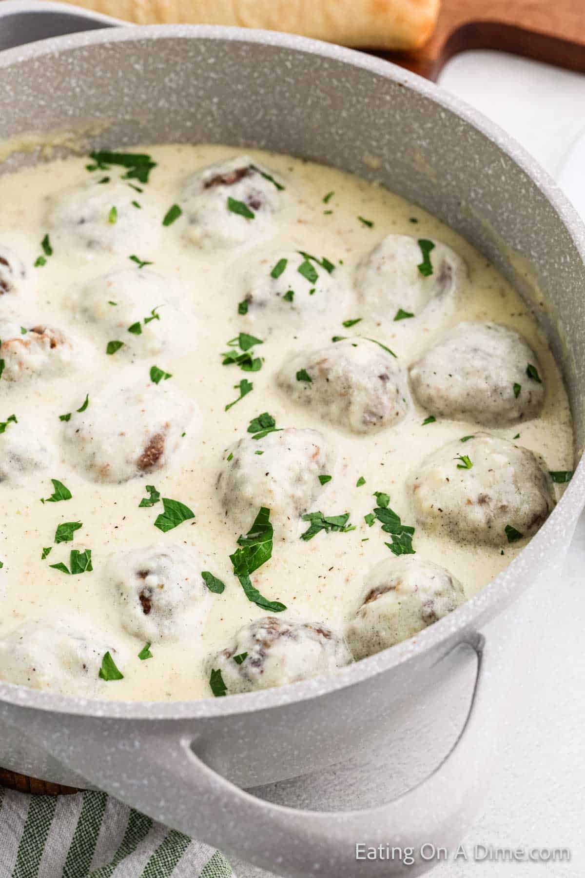 A large pot filled with creamy Alfredo meatballs garnished with chopped parsley. The meatballs are submerged in a rich, white sauce. The pot is placed on a white surface next to a cutting board with a loaf of bread.