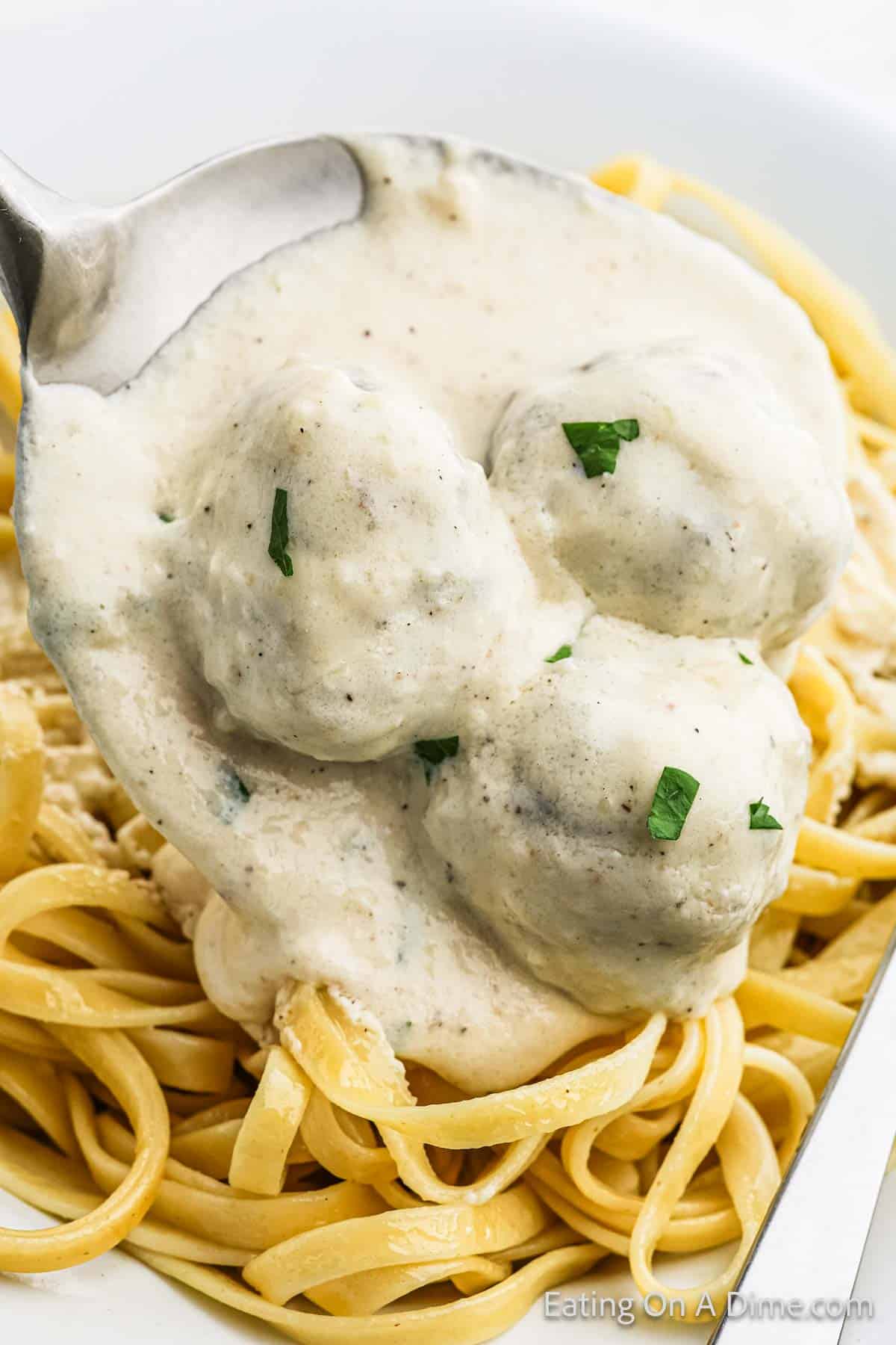 A close-up of a spoon holding three meatballs covered in a creamy Alfredo sauce, shown over a bed of fettuccine pasta. The meatballs are garnished with small parsley pieces.