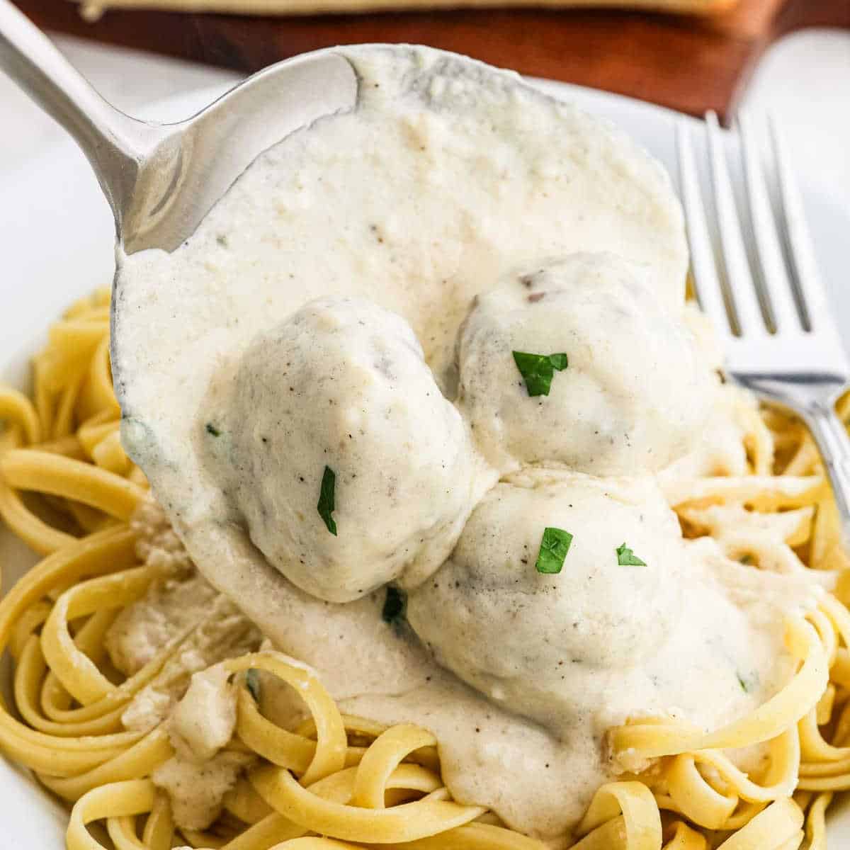 A spoon is serving creamy Swedish meatballs over a plate of cooked fettuccine pasta. The meatballs, draped in rich Alfredo sauce and garnished with small green herbs, look delectable. A fork is placed on the plate next to the pasta, completing this inviting recipe.