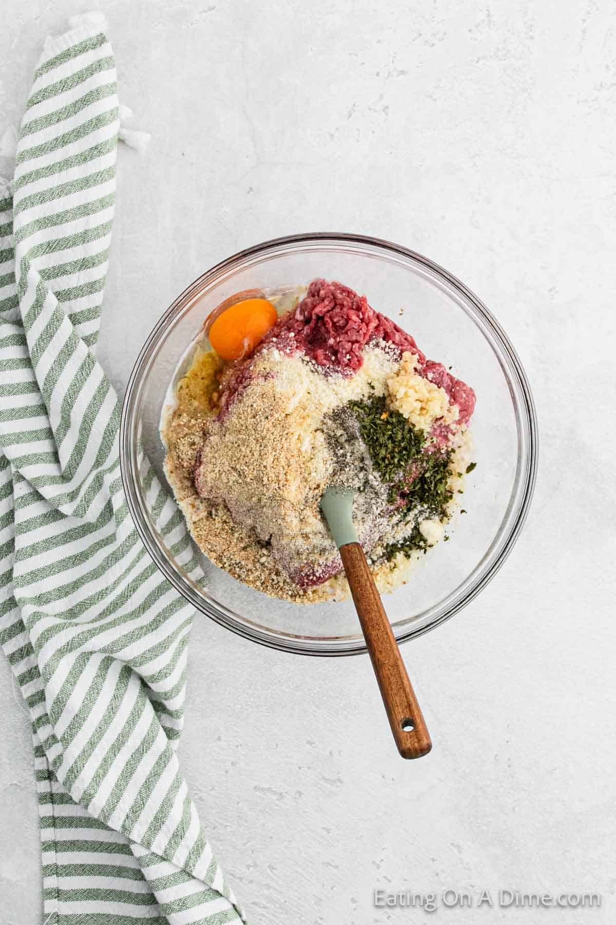 A glass bowl containing ground meat, an egg, breadcrumbs, chopped herbs, and grated cheese sets the stage for a delicious meatballs recipe. A spatula with a wooden handle rests inside the bowl. A green and white striped kitchen towel is next to the bowl on a pristine white surface.