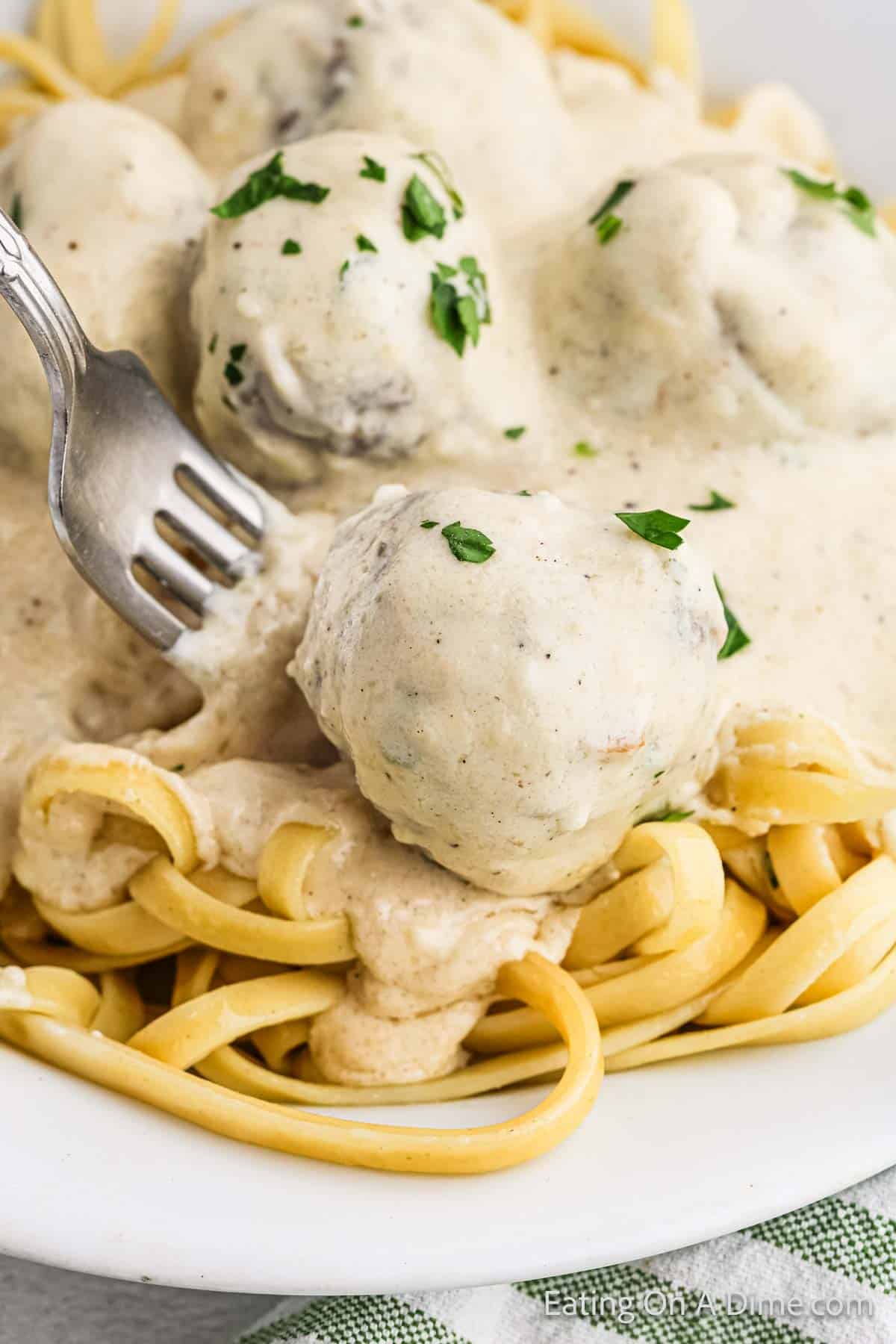 A plate of fettuccine pasta topped with savory meatballs covered in creamy Alfredo sauce. A fork is piercing one of the meatballs, and parsley garnishes the dish. The edges of a green and white checkered napkin are visible in the corner.