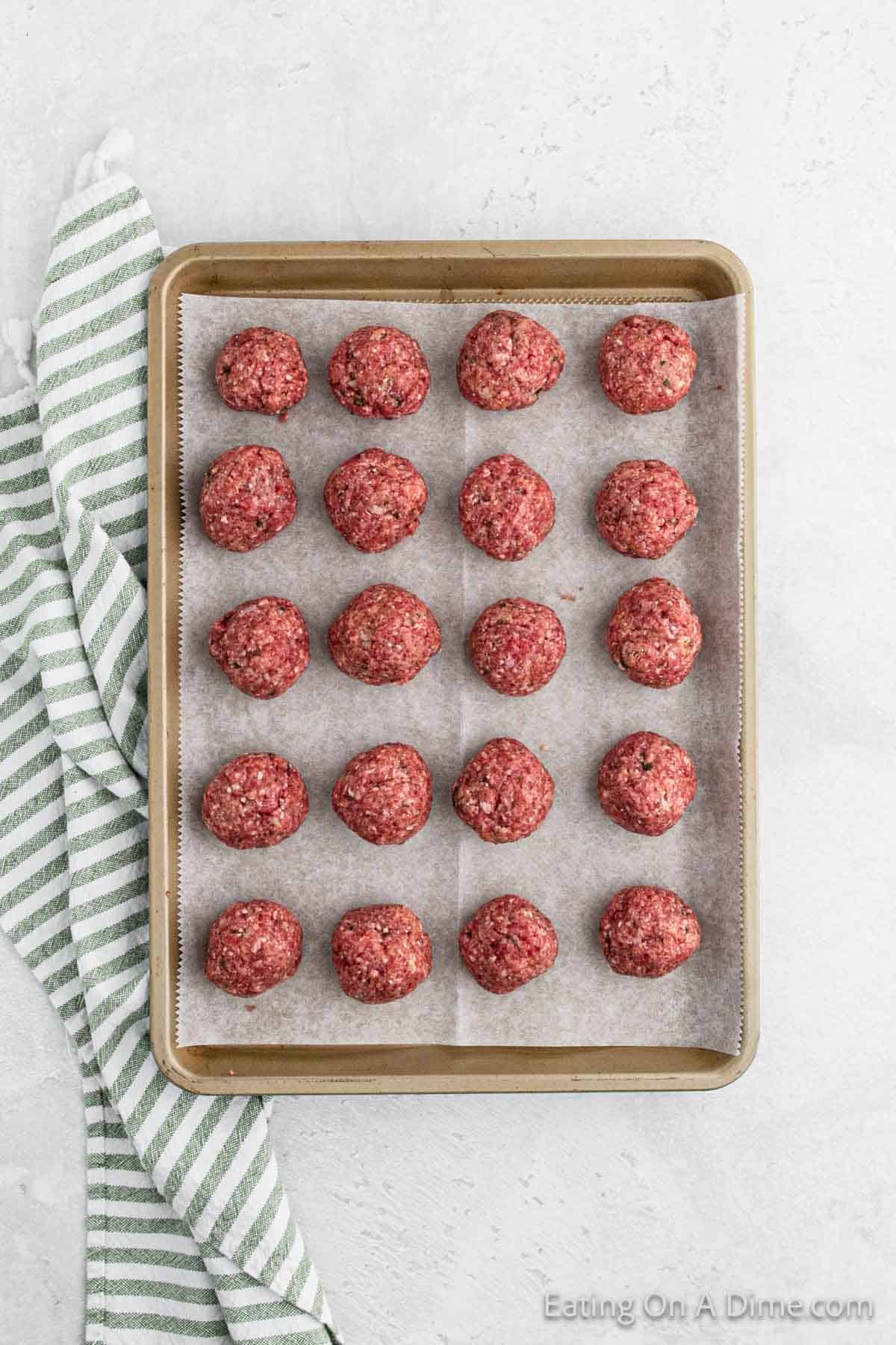 A baking sheet lined with parchment paper holds 20 raw meatballs, evenly spaced in a 4x5 grid. A green and white striped kitchen towel is draped beside the baking sheet on a light-colored countertop, ready for an Alfredo dish. The bottom right corner has the text "Eating On A Dime.com".