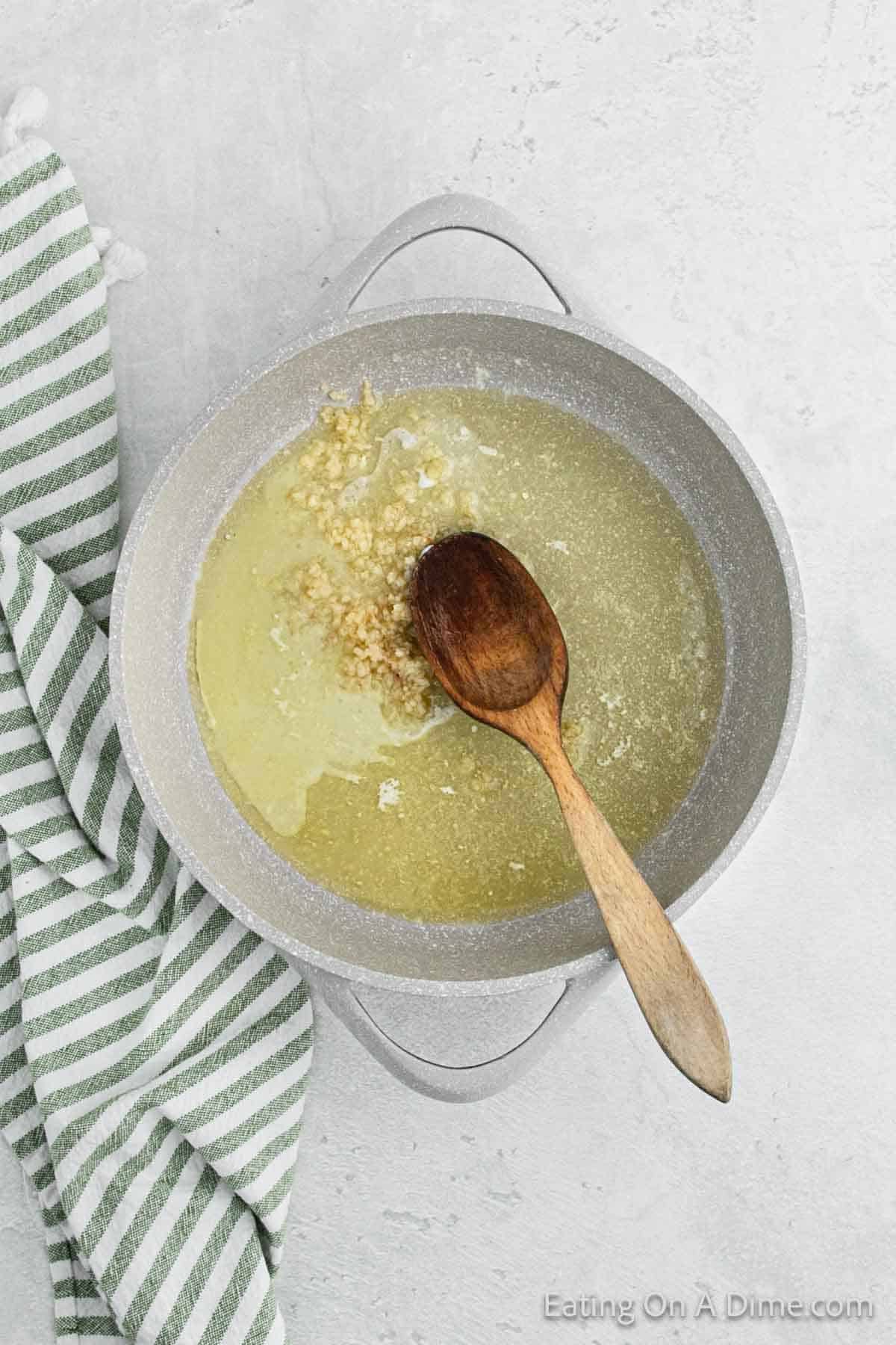 A cooking pot bubbles with a mixture of minced garlic and butter, hinting at a delectable Alfredo sauce in the making. A wooden spoon rests atop the pot. To the left, a green and white striped kitchen towel is partially visible on a grey, textured background.