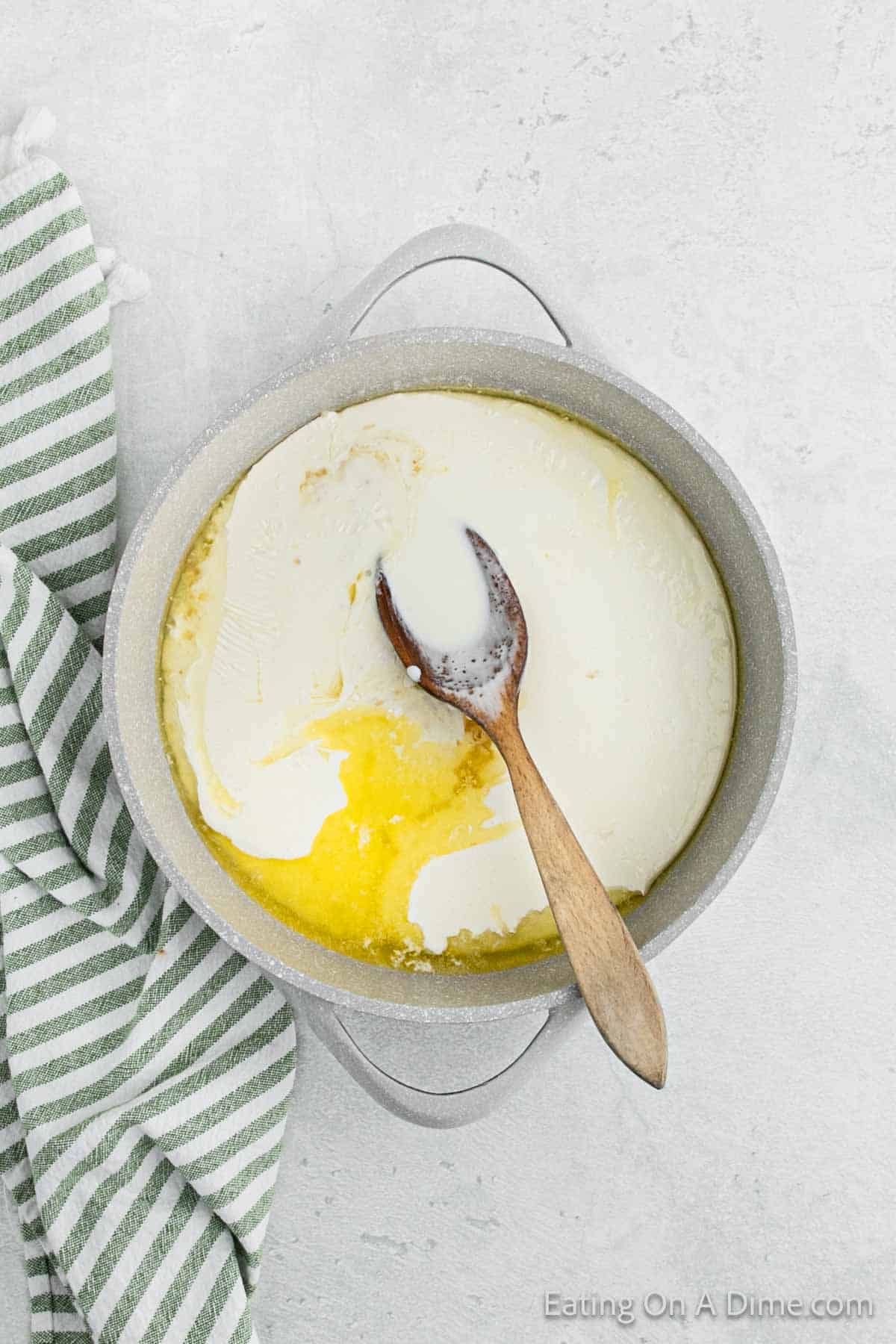 A pot on a light-colored surface contains a creamy Alfredo mixture with a layer of melted butter on top. A wooden spoon rests in the pot, partially submerged in the sauce. Nearby, a green and white striped cloth is placed next to the pot, hinting at delicious recipe preparations.