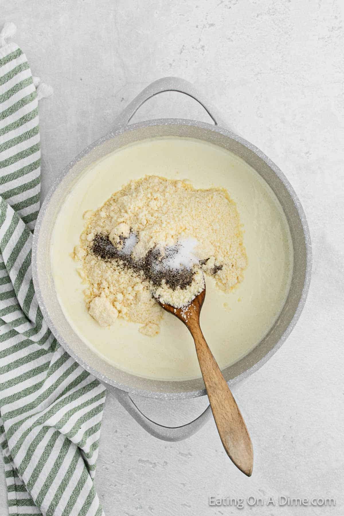 A pot filled with creamy Alfredo sauce is seen with a wooden spoon resting inside. The sauce has grated cheese and seasonings on top. A green and white striped kitchen towel is placed next to the pot on a light-colored surface.