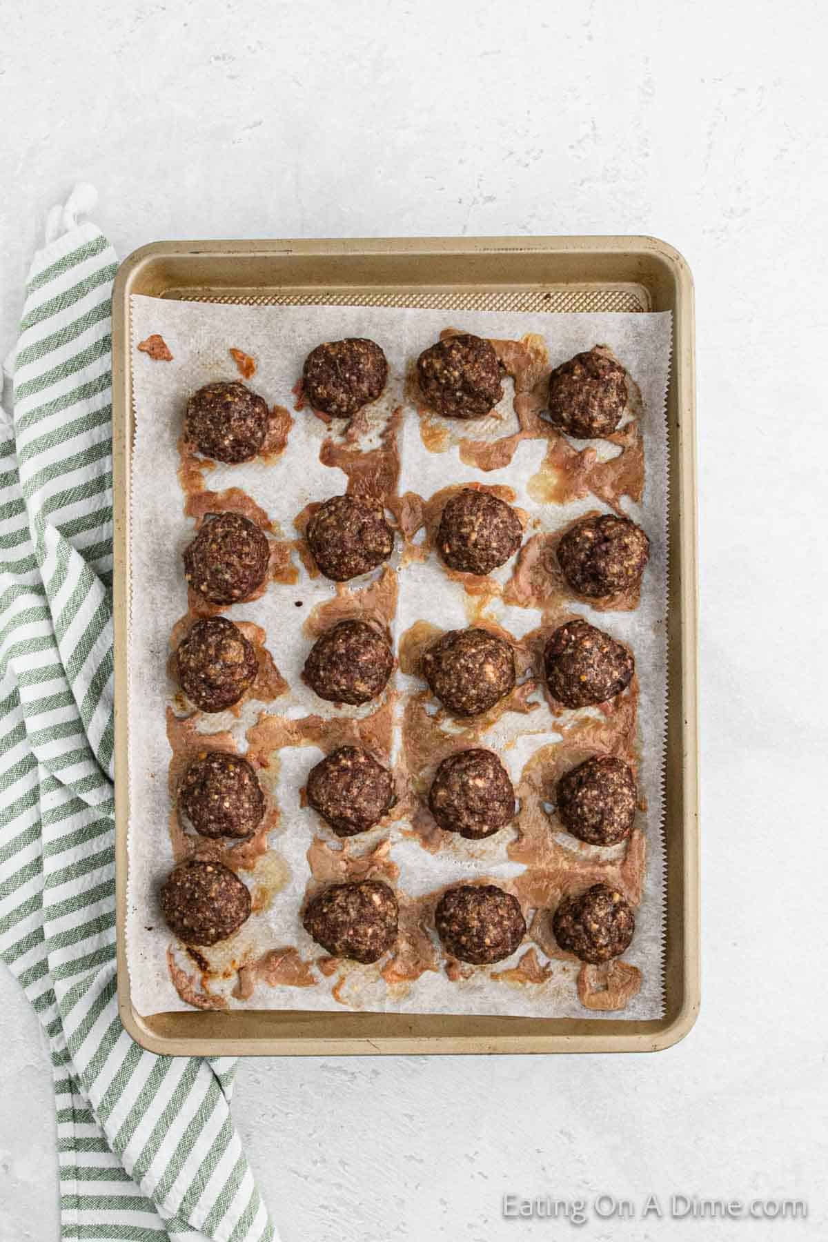 A baking tray lined with parchment paper holds 20 baked meatballs arranged in a grid. Melted cheese has oozed out around the meatballs, reminiscent of a hearty Alfredo recipe. A green and white striped cloth is placed next to the tray on the left side. The background is a light-colored surface.