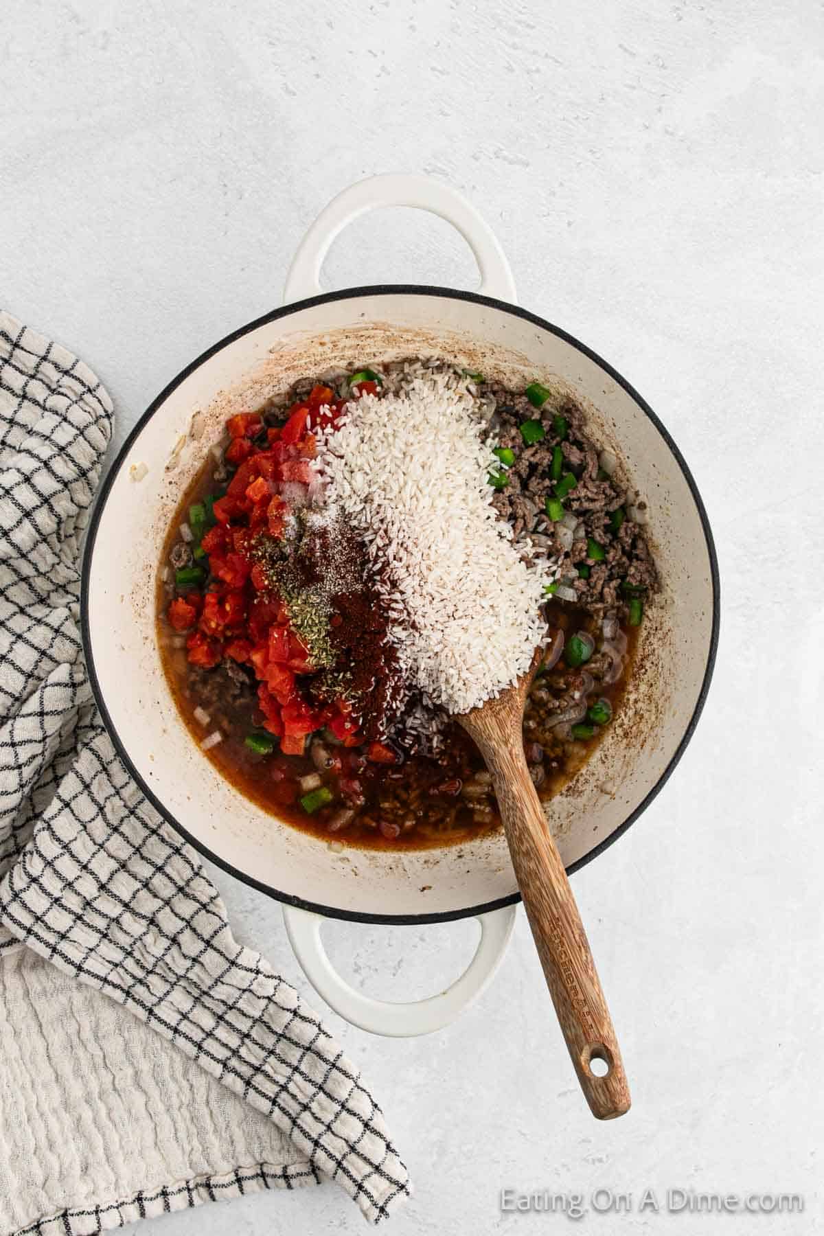 White rice, broth, seasoning added to the ground beef mixture in a skillet with a wooden spoon