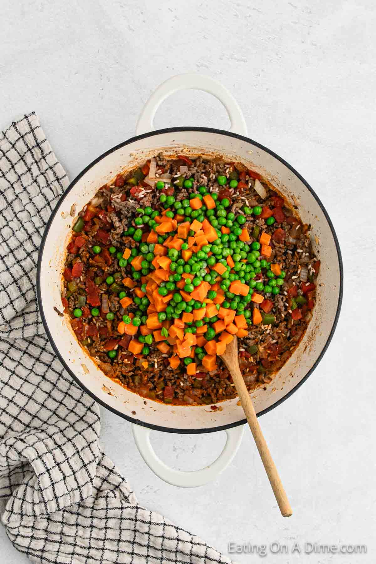 Ground beef mixture in a large pot topped with peas and carrots with a wooden spoon