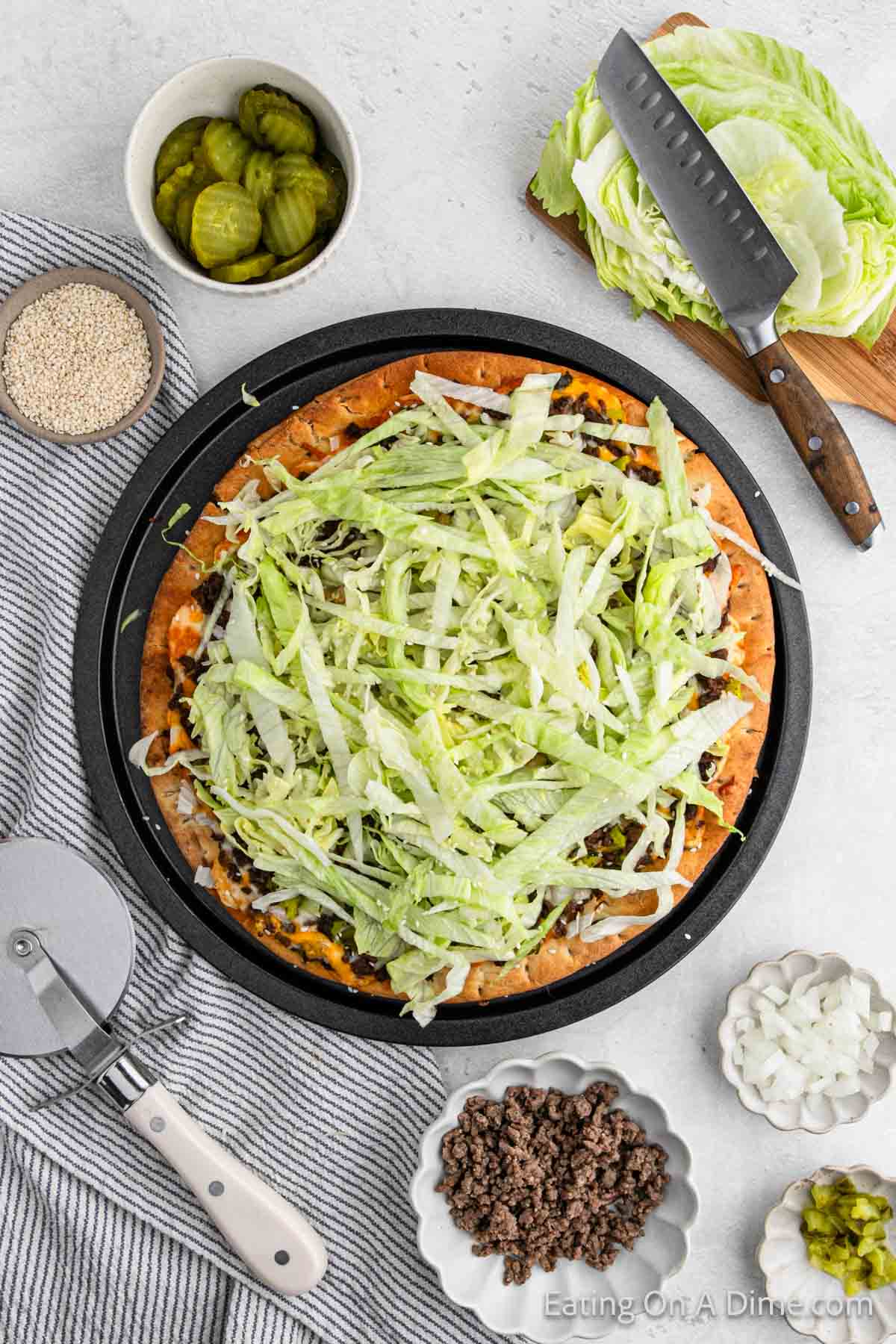 Big Mac Pizza on a pizza pan with small bowls of ground beef, chopped onions, pickles, and sesame seeds