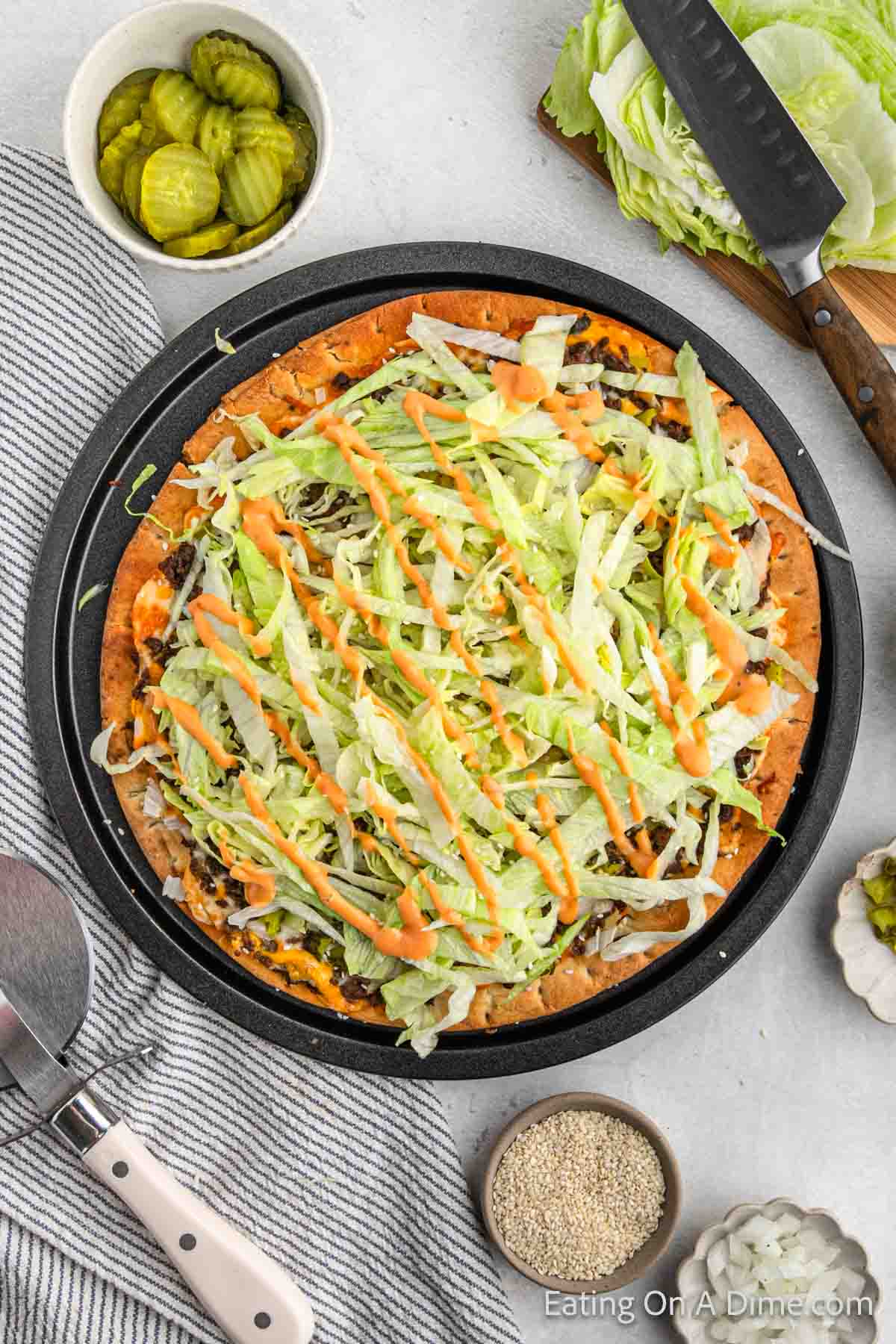 Big mac Pizza on a pizza pan topped with shredded lettuce and dressing with a bowl of pickle slices and a cutting board of lettuce