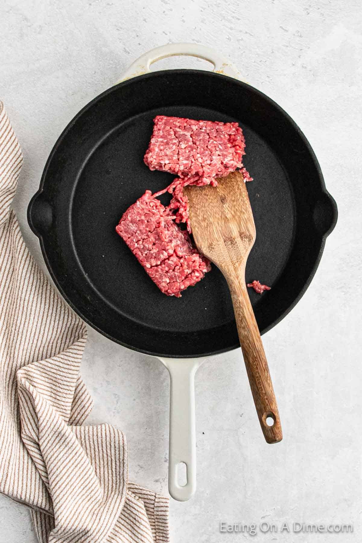 A white frying pan contains two portions of raw ground beef being browned with a wooden spatula. The pan is on a light gray surface with a beige and white striped cloth placed to the side, preparing the perfect base for a Tex-Mex Cowboy Queso dip.