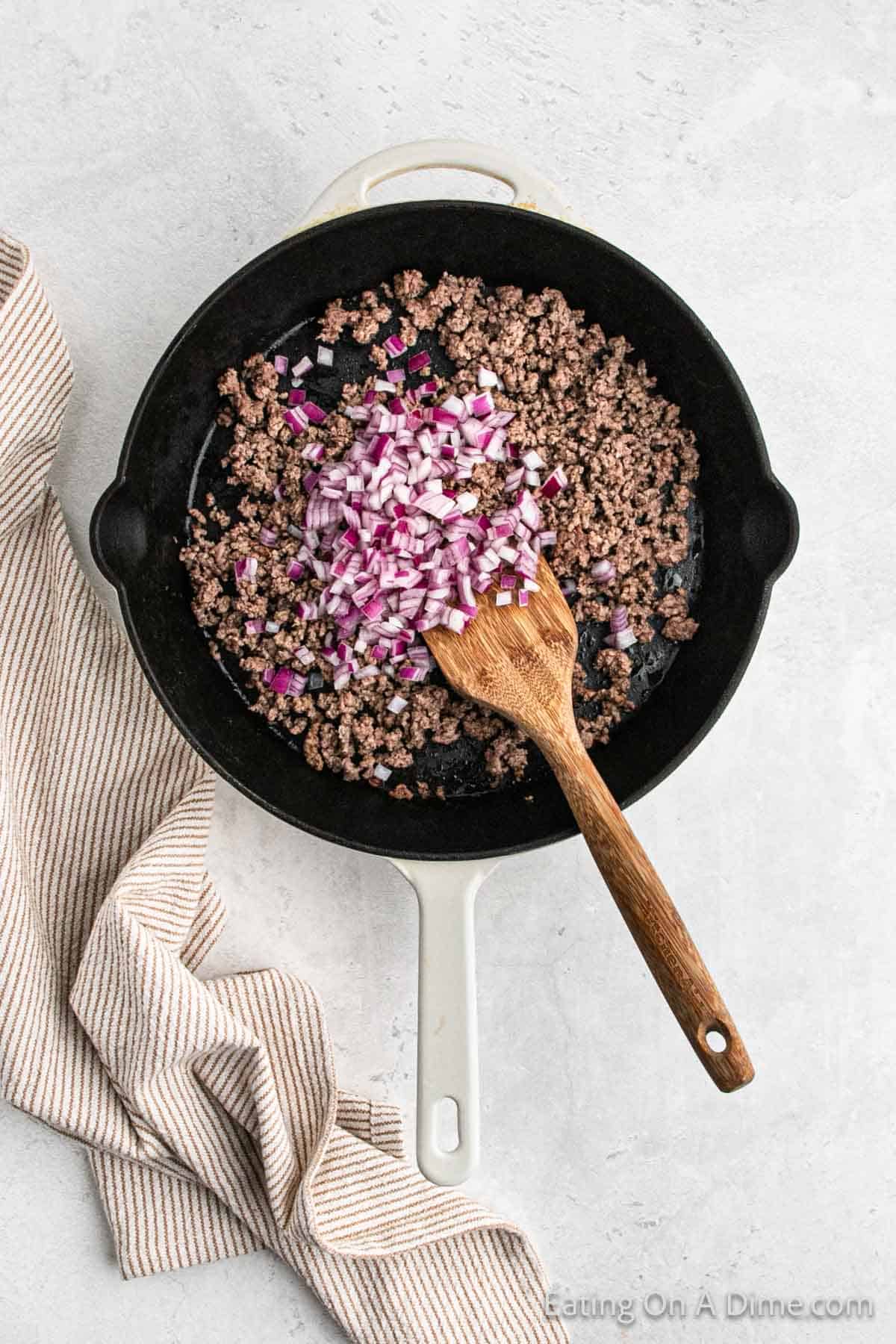 A white cast iron skillet containing ground beef and diced red onions being stirred with a wooden spatula, ready for a hearty Cowboy Queso. The skillet rests on a light gray surface, with a striped kitchen towel partially visible beside it.