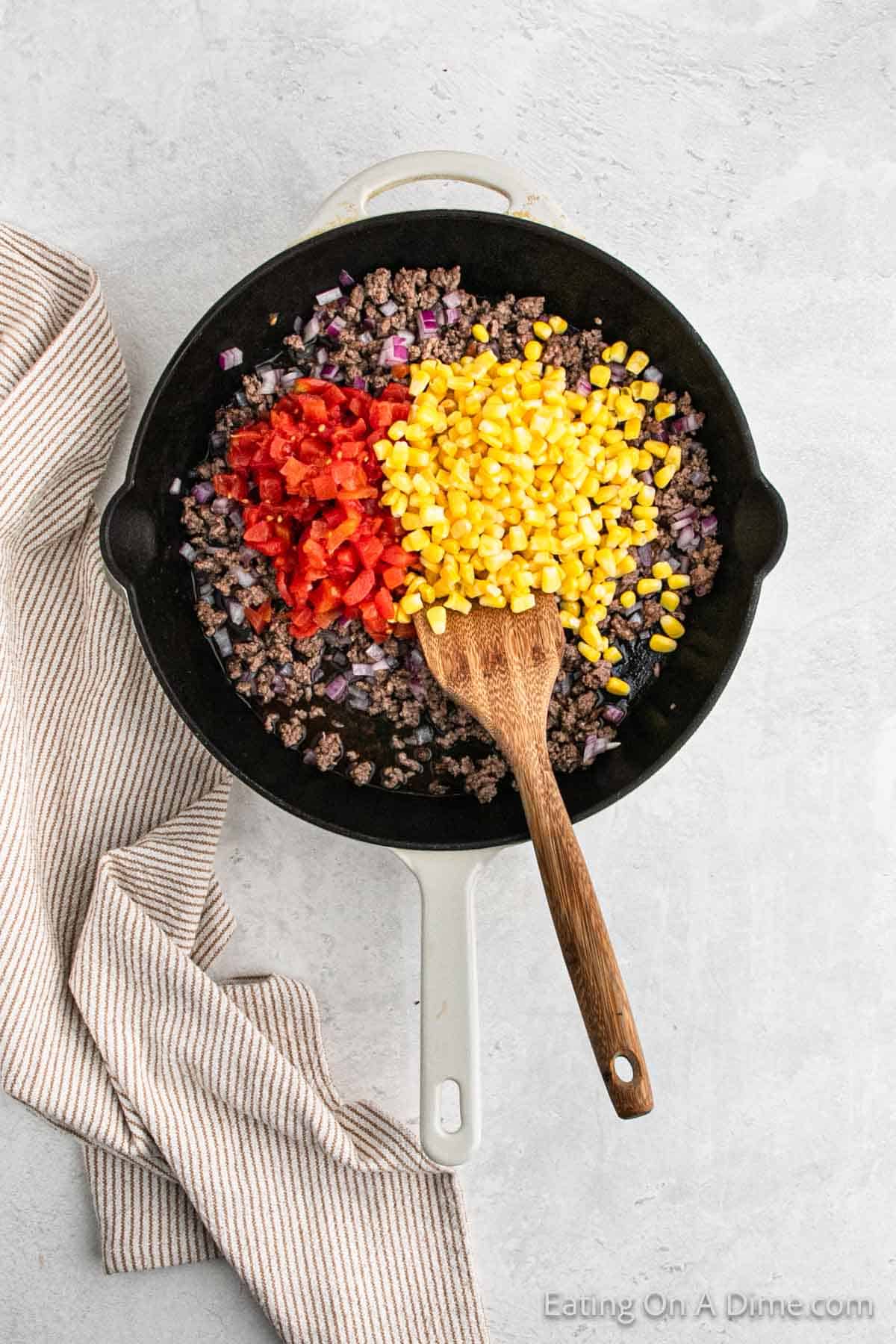 Corn, diced tomatoes topped ground beef in a skillet with a wooden spoon