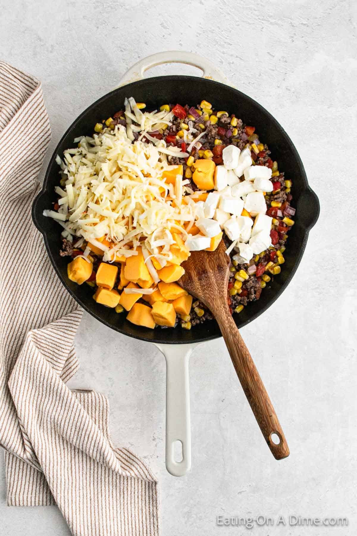 Ground sausage mixture topped with diced cream cheese, diced velveeta and shredded cheese in a skillet with a wooden spoon