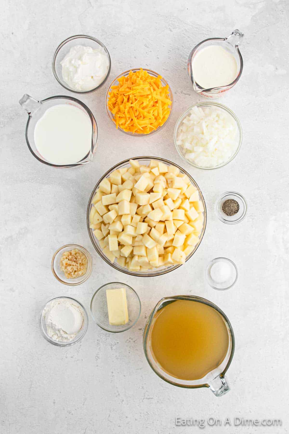 A top-down view of various ingredients arranged on a light surface, resembling the perfect setup for a baked potato soup in a crock pot, including cubed potatoes, grated cheddar cheese, chopped onions, chicken broth, milk, heavy cream, sour cream, butter, garlic, flour, and ground pepper.