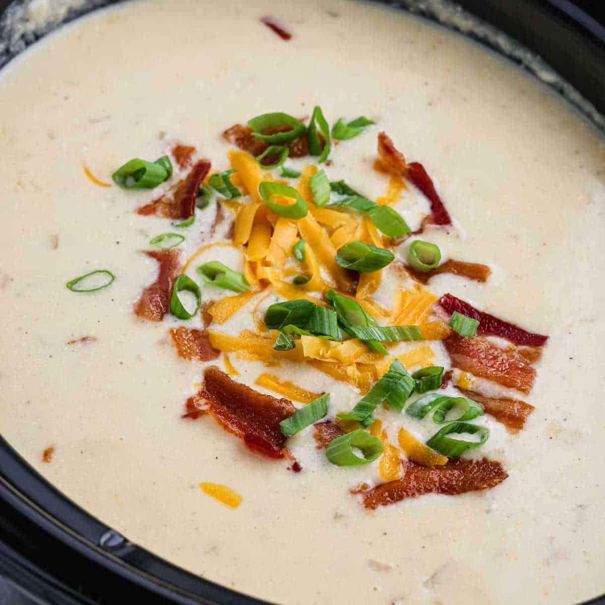 A close-up of a bowl of creamy baked potato soup garnished with chopped green onions, shredded cheese, and crispy bacon bits. The rich, thick soup is beige in color and appears seasoned with visible herbs and pepper, offering a comforting aroma straight from the crock pot.