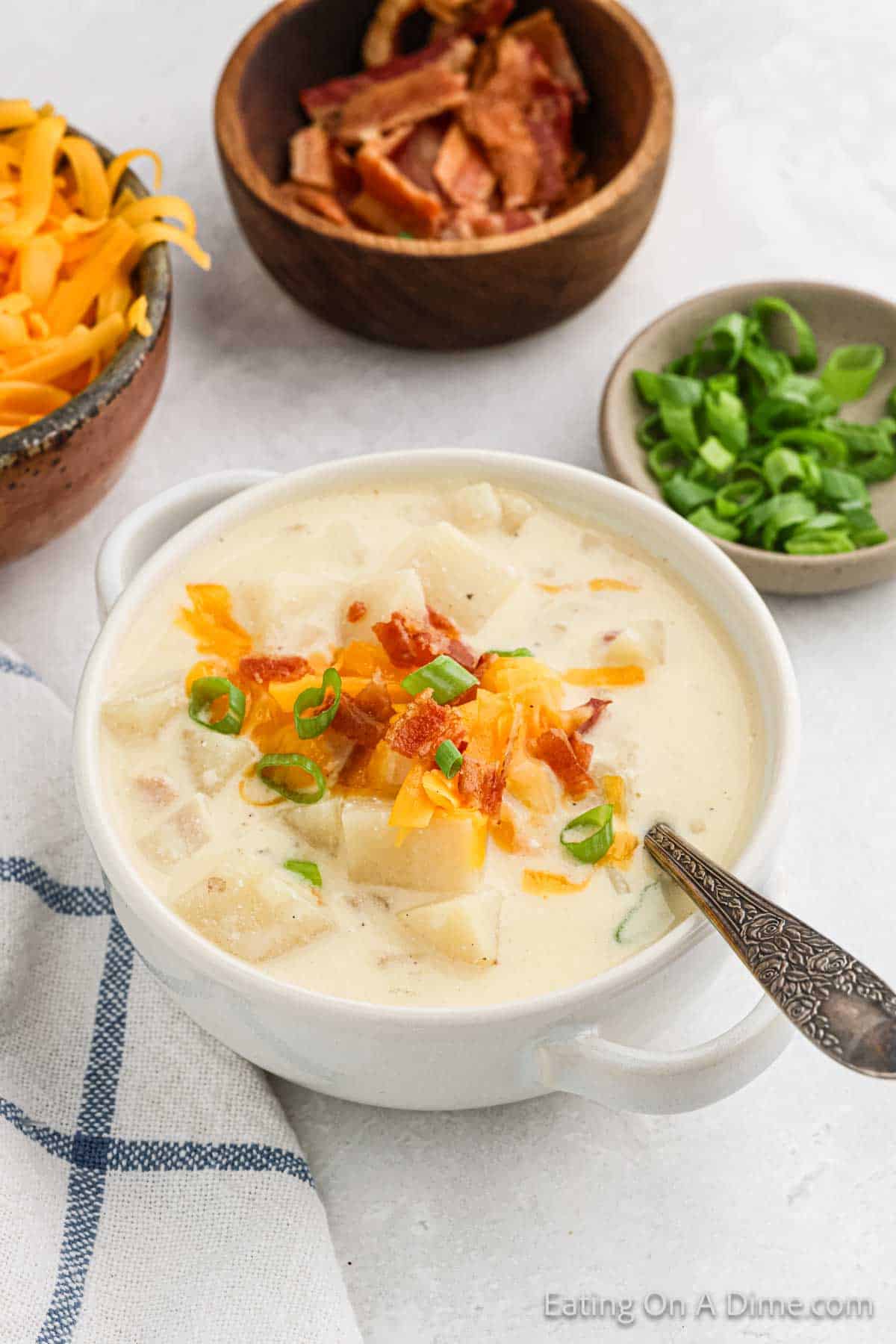 Creamy baked potato soup in a white bowl, topped with shredded cheese, bacon bits, and sliced green onions. A spoon is in the bowl. Surrounding it are small dishes containing additional toppings like more shredded cheese, bacon, and green onions—all simmered to perfection in a Crock Pot.