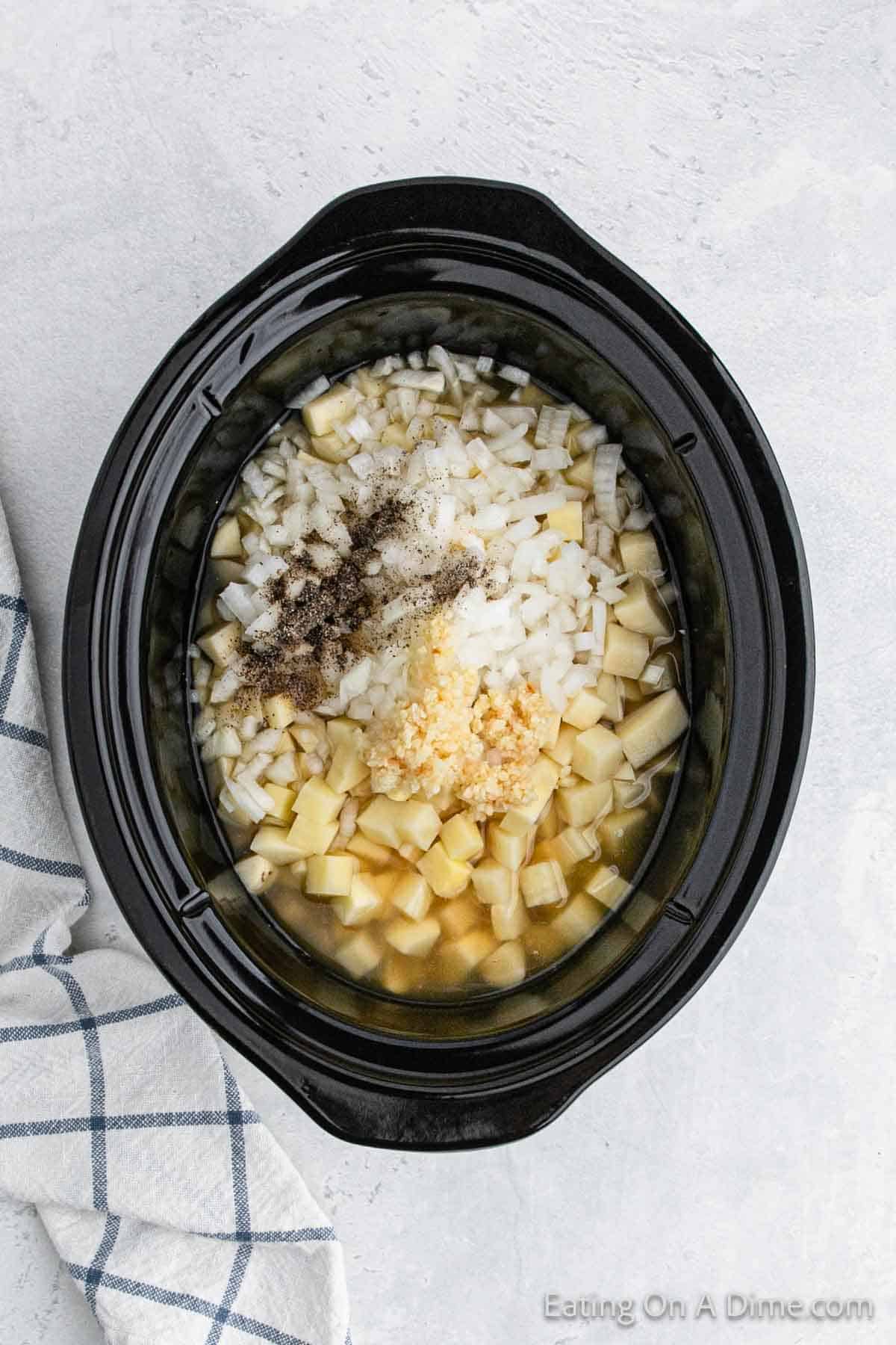 A Crock Pot filled with chopped potatoes, diced onions, minced garlic, and ground pepper simmers to create a hearty Baked Potato Soup. A checkered kitchen towel is partially visible on the left side of the image.