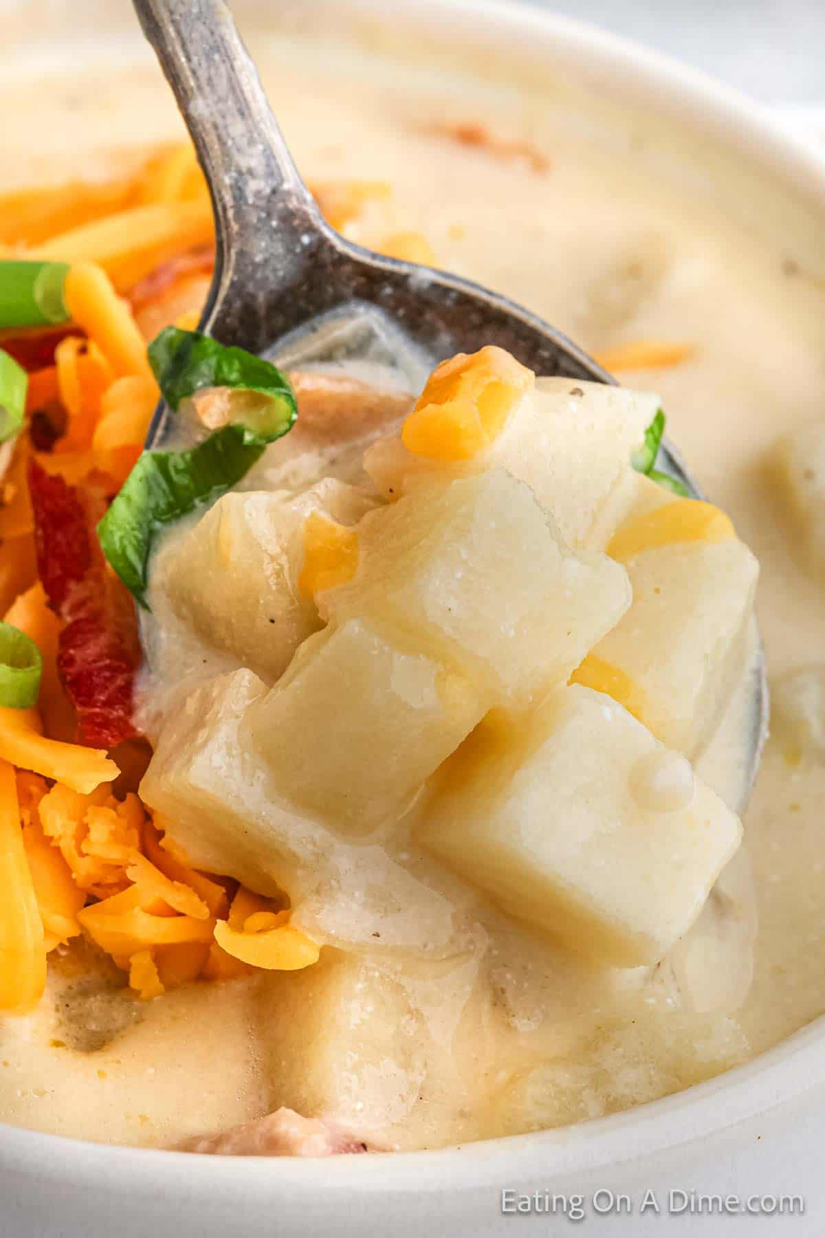 A close-up of a spoonful of Baked Potato Soup. The spoon holds chunks of potato garnished with shredded cheddar cheese and chopped green onions. Made in a Crock Pot, the soup appears thick and rich, with a velvety texture.