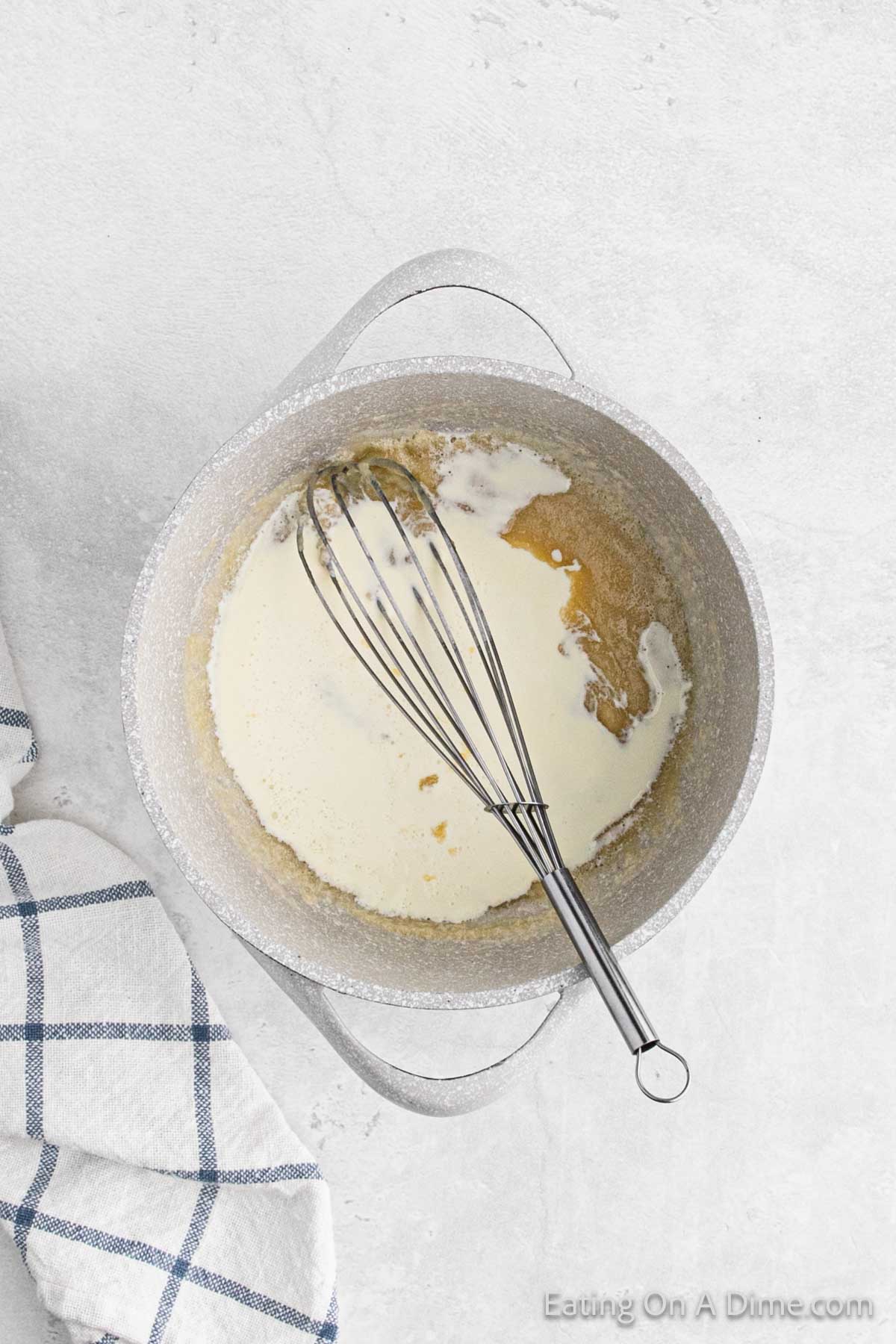 A top-down view of a metal pot on a light surface with a whisk inside. The pot holds the beginnings of baked potato soup, with a light cream being blended into a darker mixture. A white and blue checkered towel is partially visible on the left side of the image, adding to the cozy kitchen vibe.