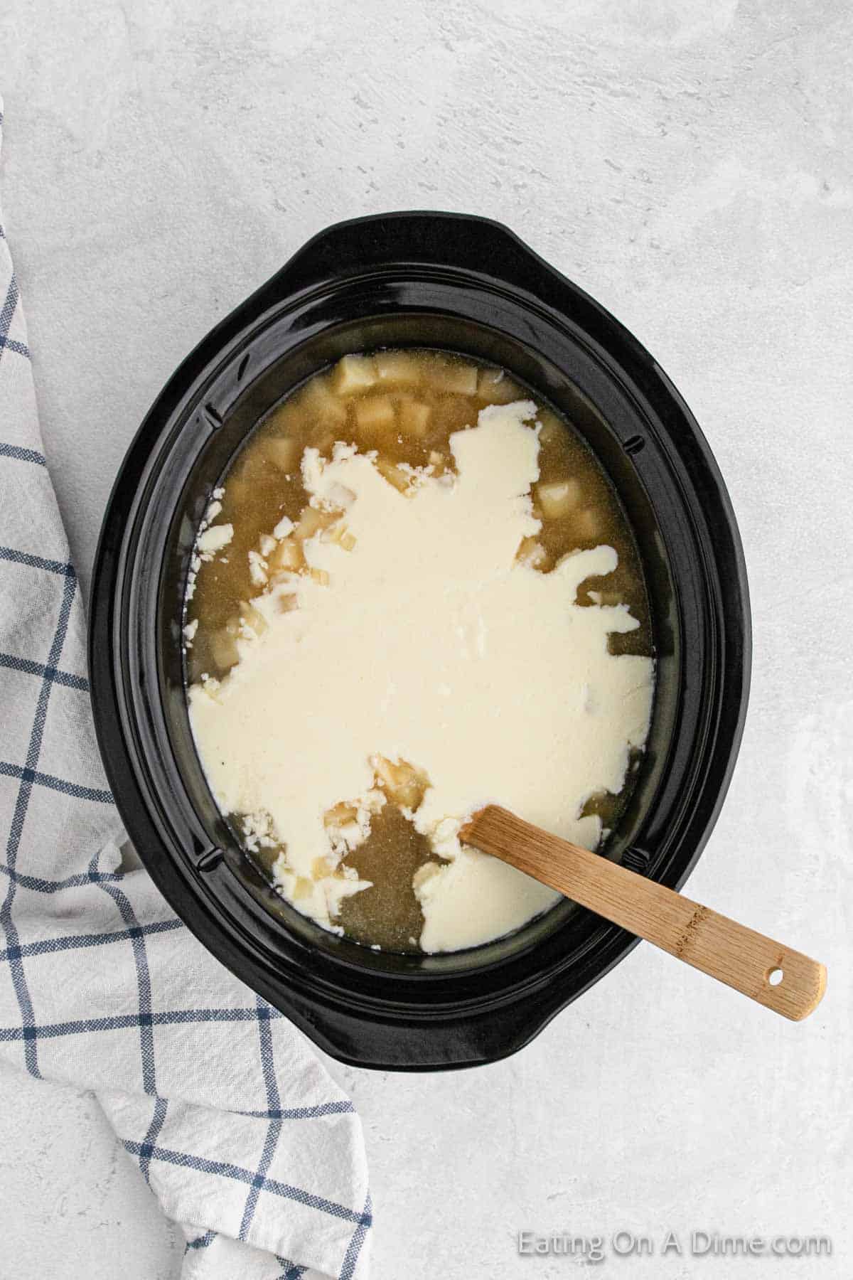 A Crock Pot filled with creamy baked potato soup in the making. Visible are chunks of vegetables and a wooden spoon stirring the mixture. A blue and white checkered kitchen towel is placed beside the slow cooker on a light-colored countertop.