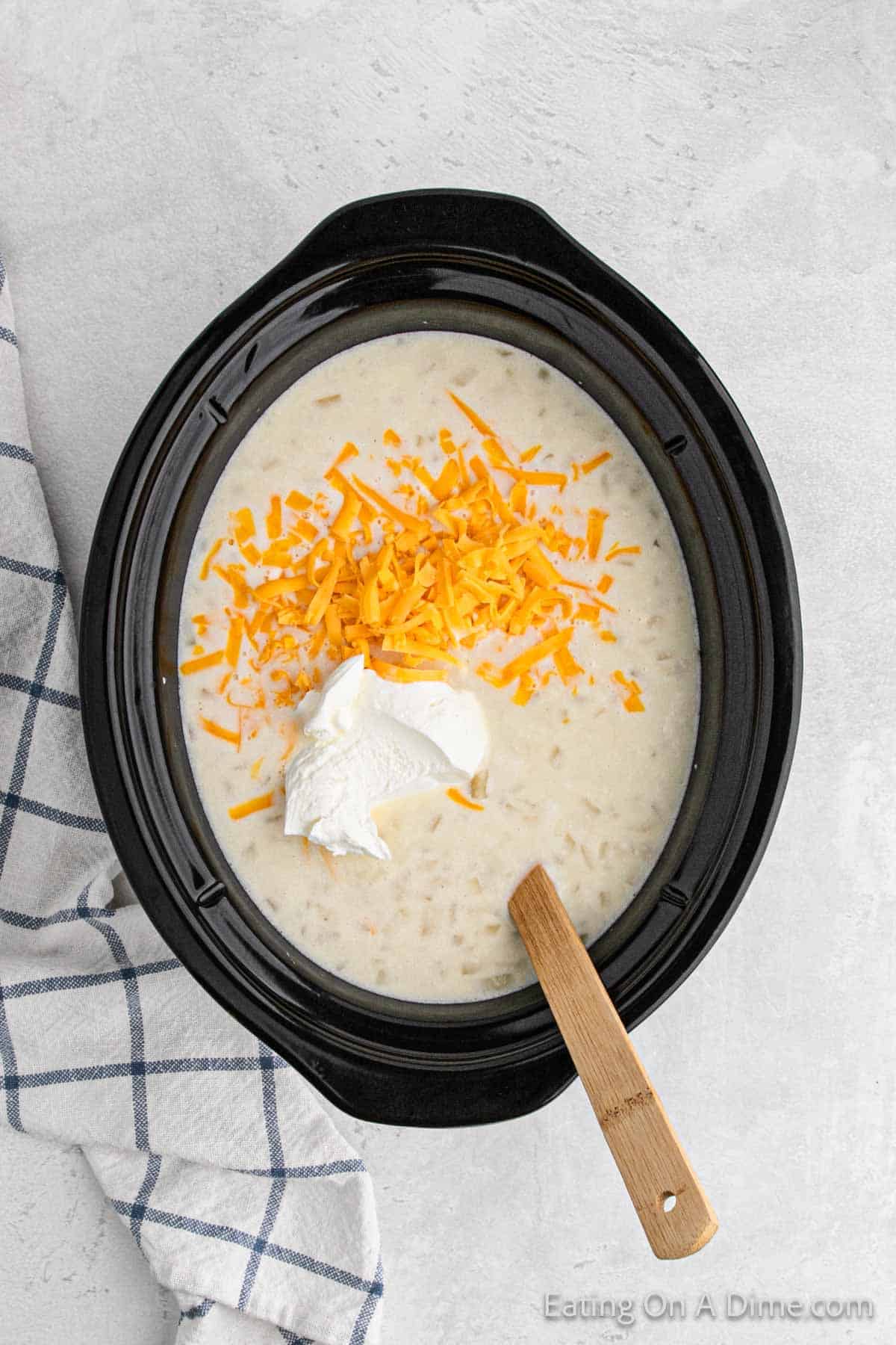 A black Crock Pot is filled with creamy Baked Potato Soup, topped with a dollop of sour cream and shredded cheddar cheese. A wooden spoon rests in the soup. A light checkered cloth lies next to the Crock Pot on a white surface.