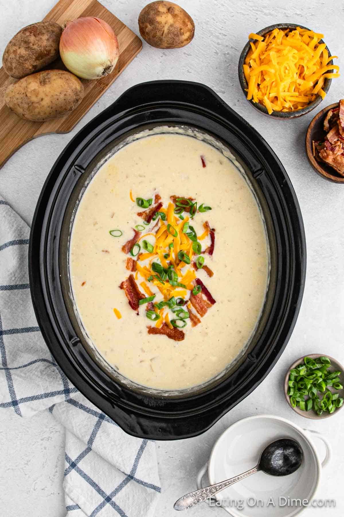 A Crock Pot filled with creamy Baked Potato Soup topped with shredded cheese, bacon bits, and chopped green onions. Surrounding the Crock Pot are bowls of shredded cheese, bacon bits, and chopped green onions, along with whole potatoes and an onion on a cutting board.