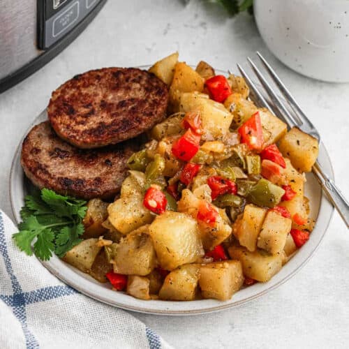 A plate of seasoned, diced breakfast potatoes mixed with red and green bell peppers and onions, garnished with cilantro, sits next to two browned sausage patties. A fork rests on the plate, with a checkered napkin beside it and a portion of a crockpot visible in the background.