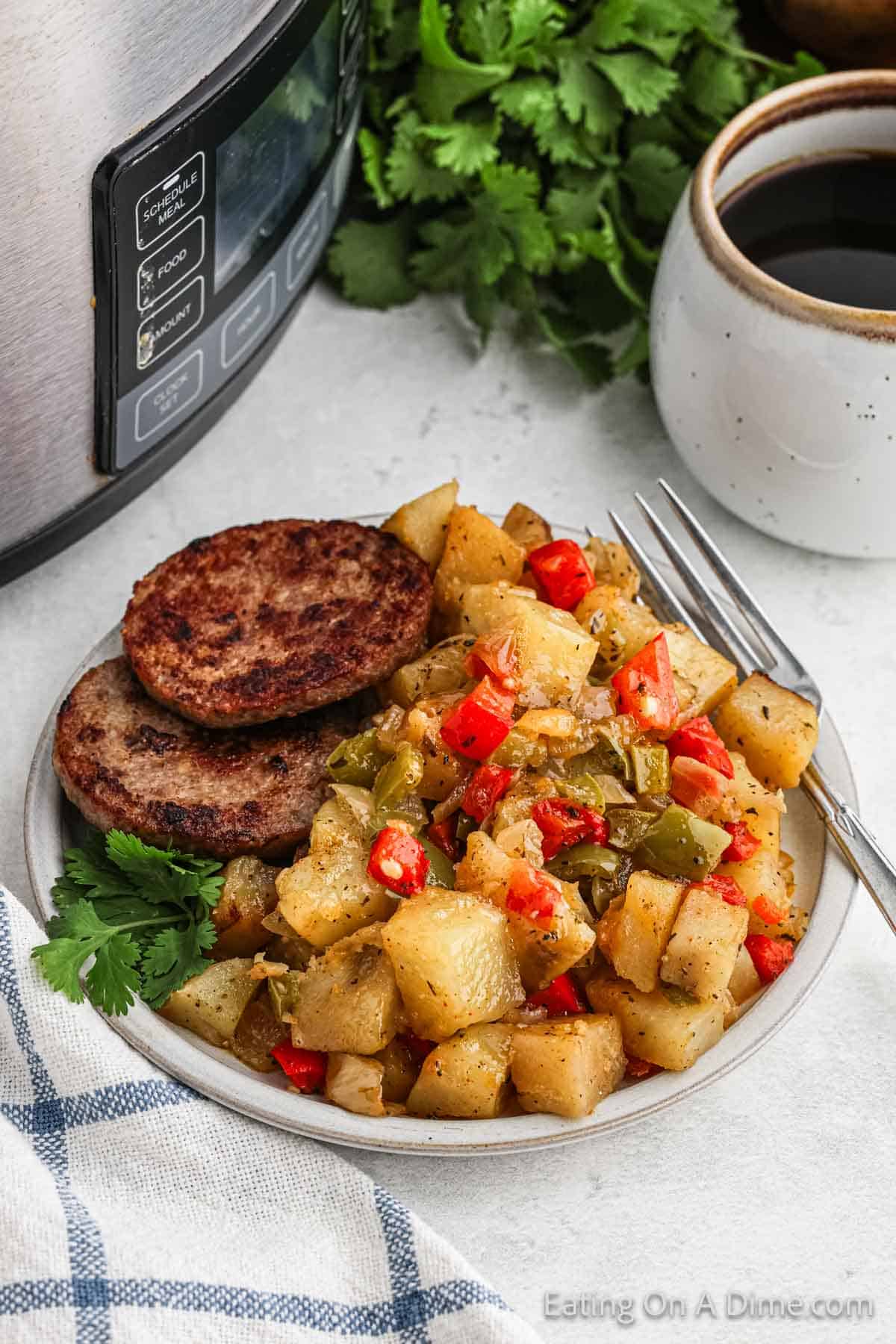 A plate of diced breakfast potatoes mixed with red and green bell peppers is served alongside two sausage patties. The dish sits next to a crockpot, with a cup of coffee and some fresh herbs in the background. A blue and white cloth is partially visible.