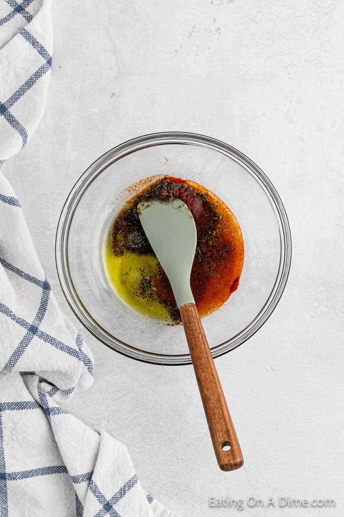 A clear glass bowl contains a mixture of olive oil, spices, and seasoning, with a grey silicone spatula resting inside. The bowl is placed on a light grey surface next to a white and blue checkered cloth napkin. The corner of the image has text that says: "Eating On A Dime.com", hinting at tasty crockpot breakfast ideas.