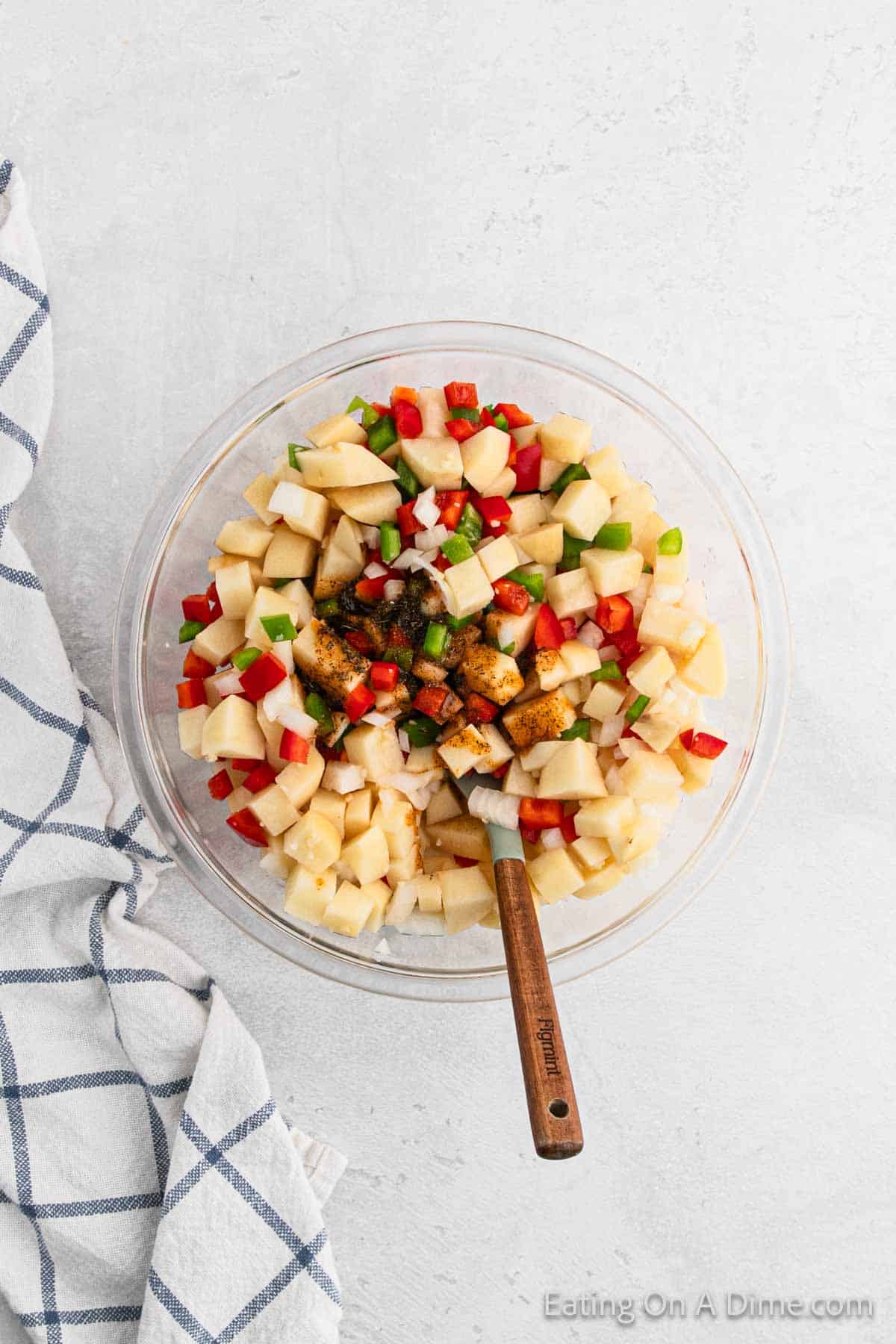 A clear glass bowl filled with chopped apples, diced red and green bell peppers, small chunks of cooked chicken, and tender potatoes, mixed with seasoning. A wooden spoon rests inside the bowl. A blue and white checkered cloth is placed beside the bowl on a light, textured surface.