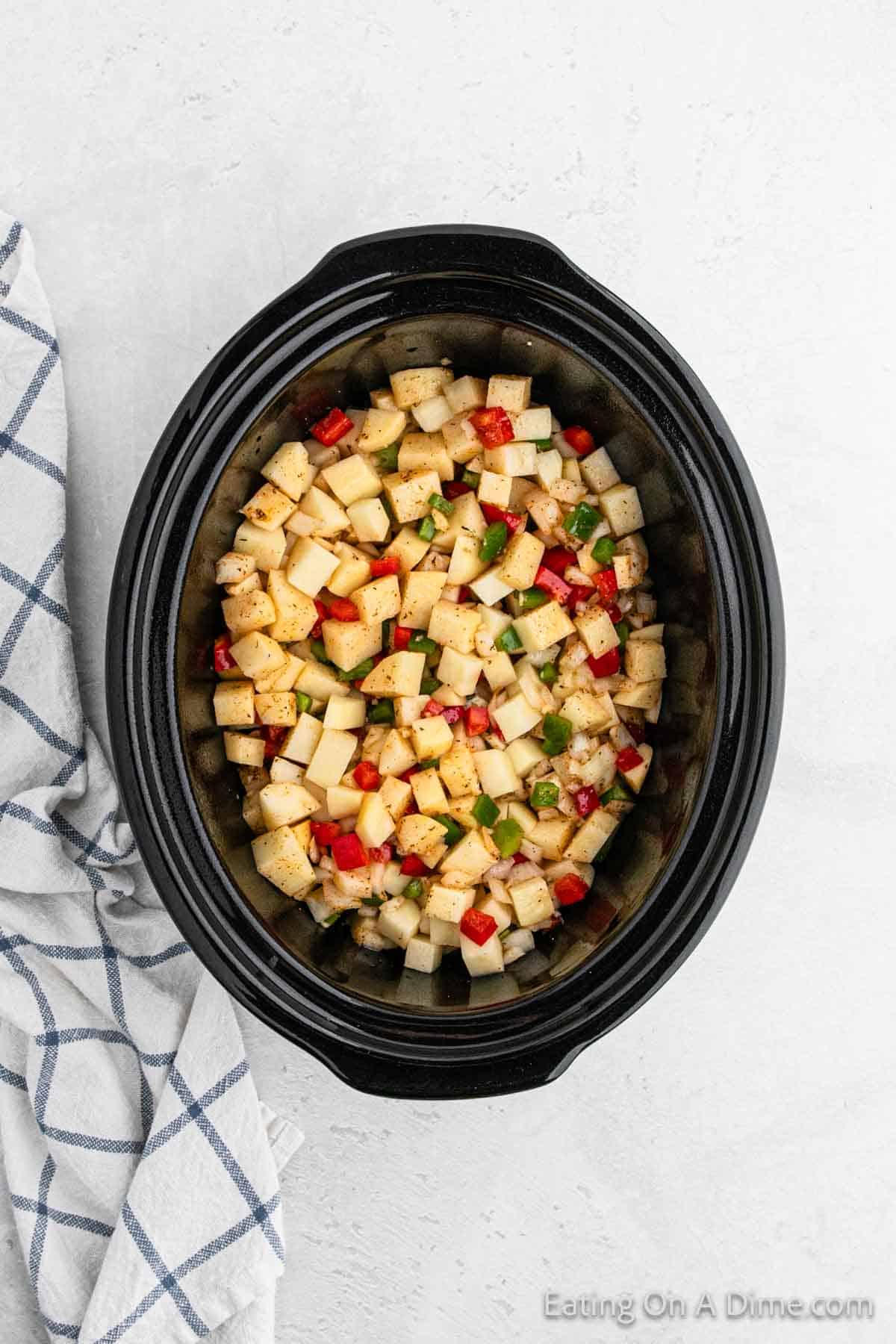 A black crockpot filled with a colorful mixture of breakfast potatoes, red bell peppers, green bell peppers, and onions. The slow cooker sits on a light countertop next to a white kitchen towel with a blue grid pattern. The image is captioned "Eating On A Dime.com".