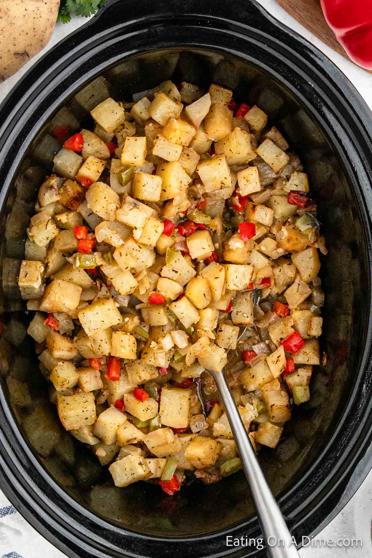 A close-up of diced potatoes mixed with red bell peppers and seasonings in a black crockpot. A silver spoon is placed in the center, ready to serve the breakfast dish. The mixture appears well-cooked, with a golden-brown color accentuated by the red peppers.