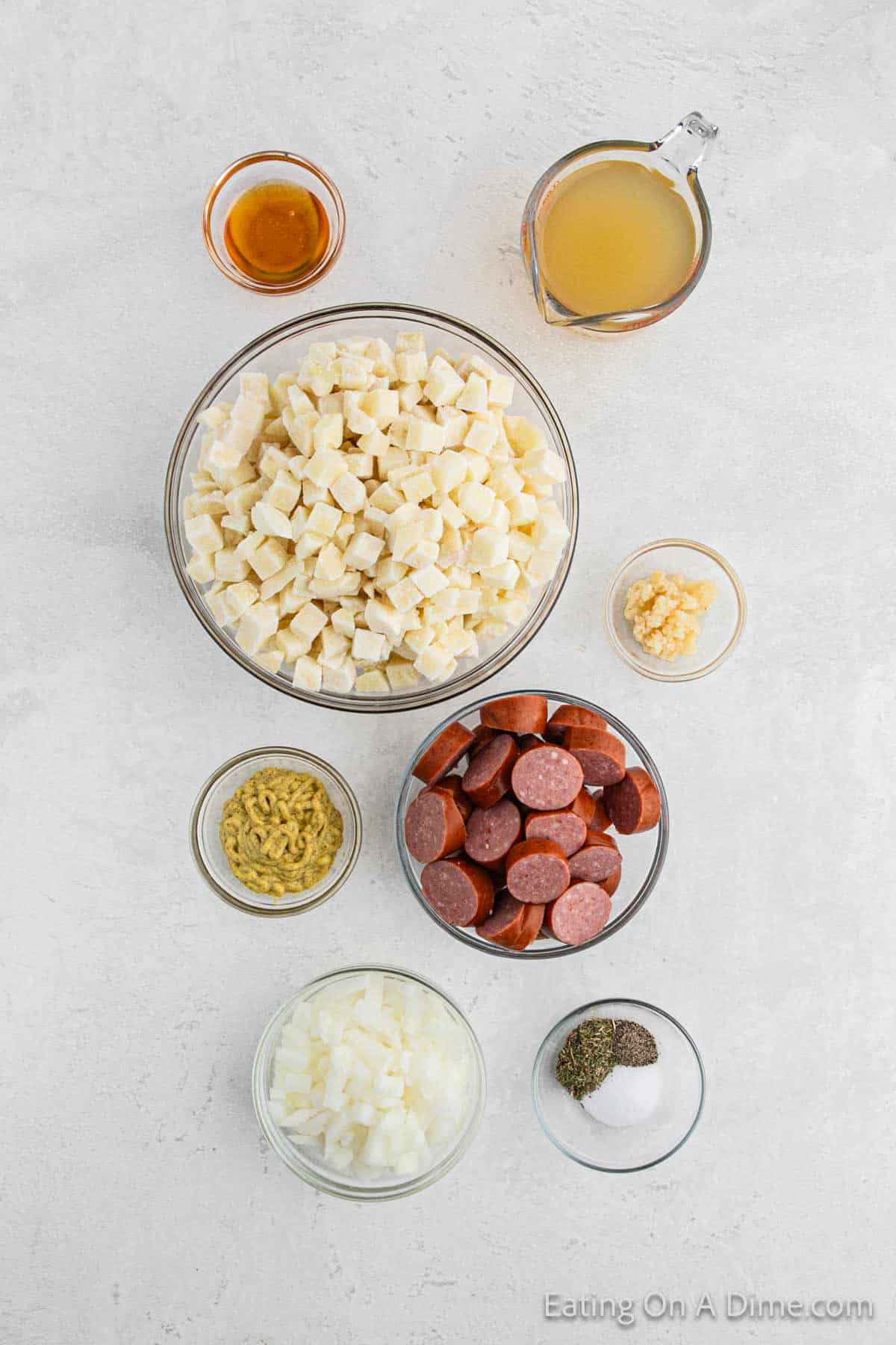 A flat lay image of ingredients for a dish. These include a bowl of diced potatoes, sliced kielbasa sausage, chopped onions, minced garlic, seasoning spices, mustard, a jug of broth for the crock pot, and a small bowl of honey.