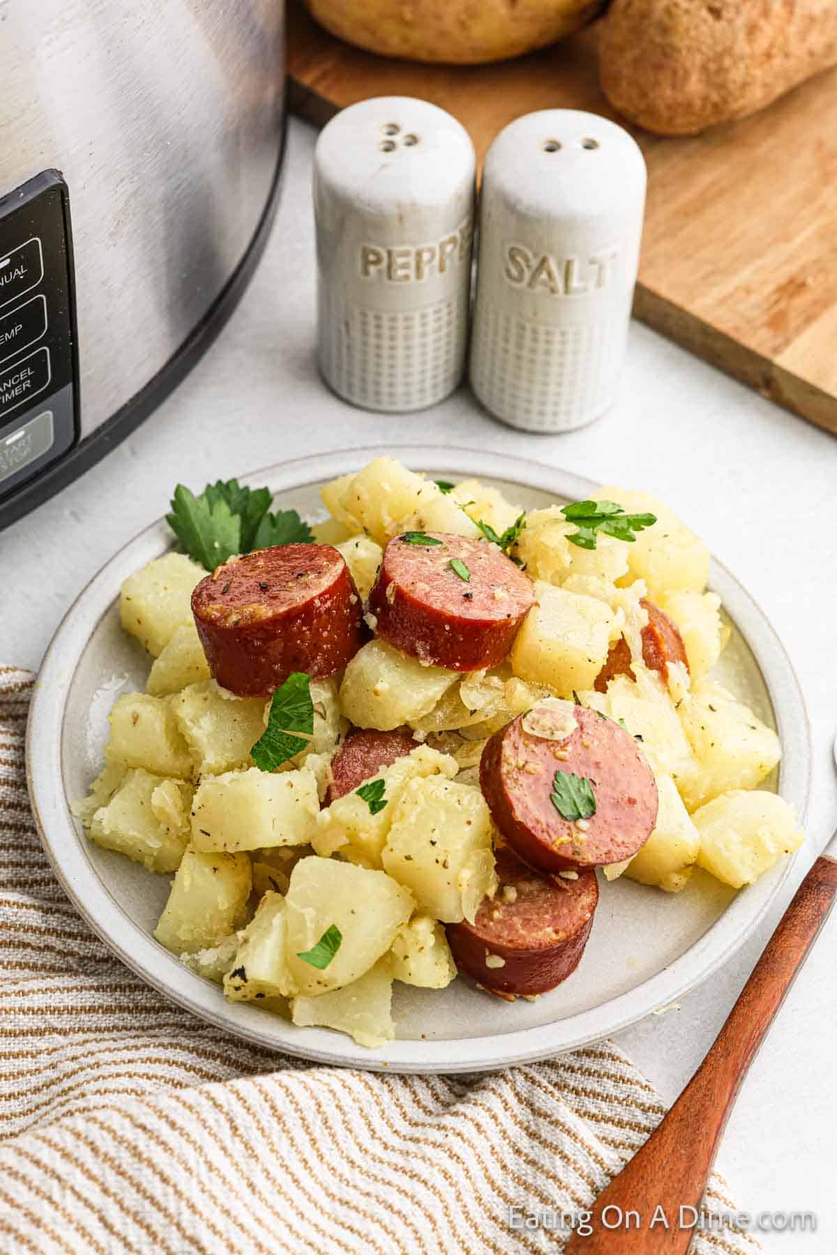 A plate of sliced kielbasa and potatoes garnished with herbs is placed next to an instant pot and a set of salt and pepper shakers. A striped cloth and a wooden spoon are positioned nearby, creating a cozy kitchen setting.