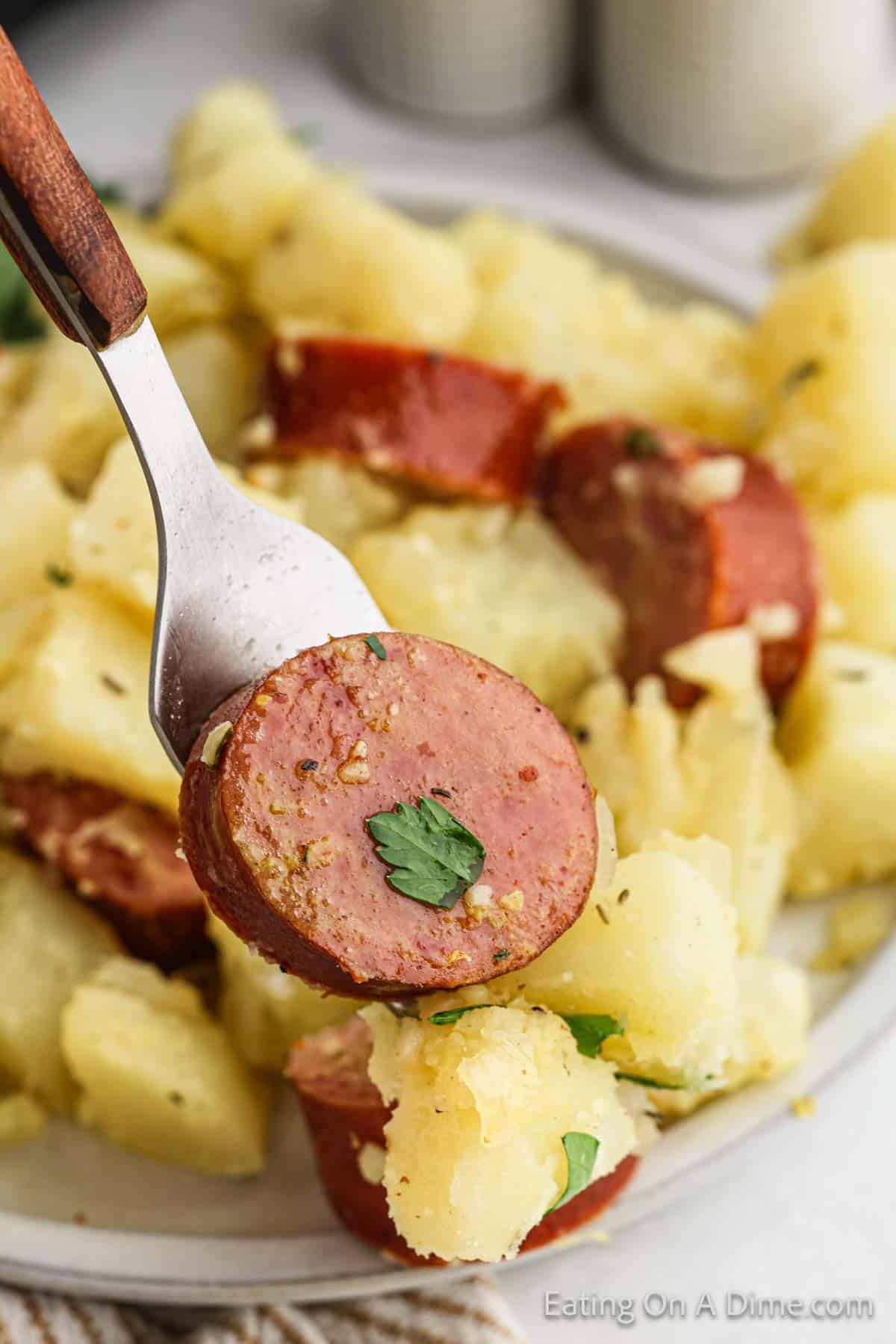 A close-up of a fork holding a piece of kielbasa above a plate of diced potatoes mixed with herbs, accompanied by other slices of sausage. The dish appears to be garnished with parsley.