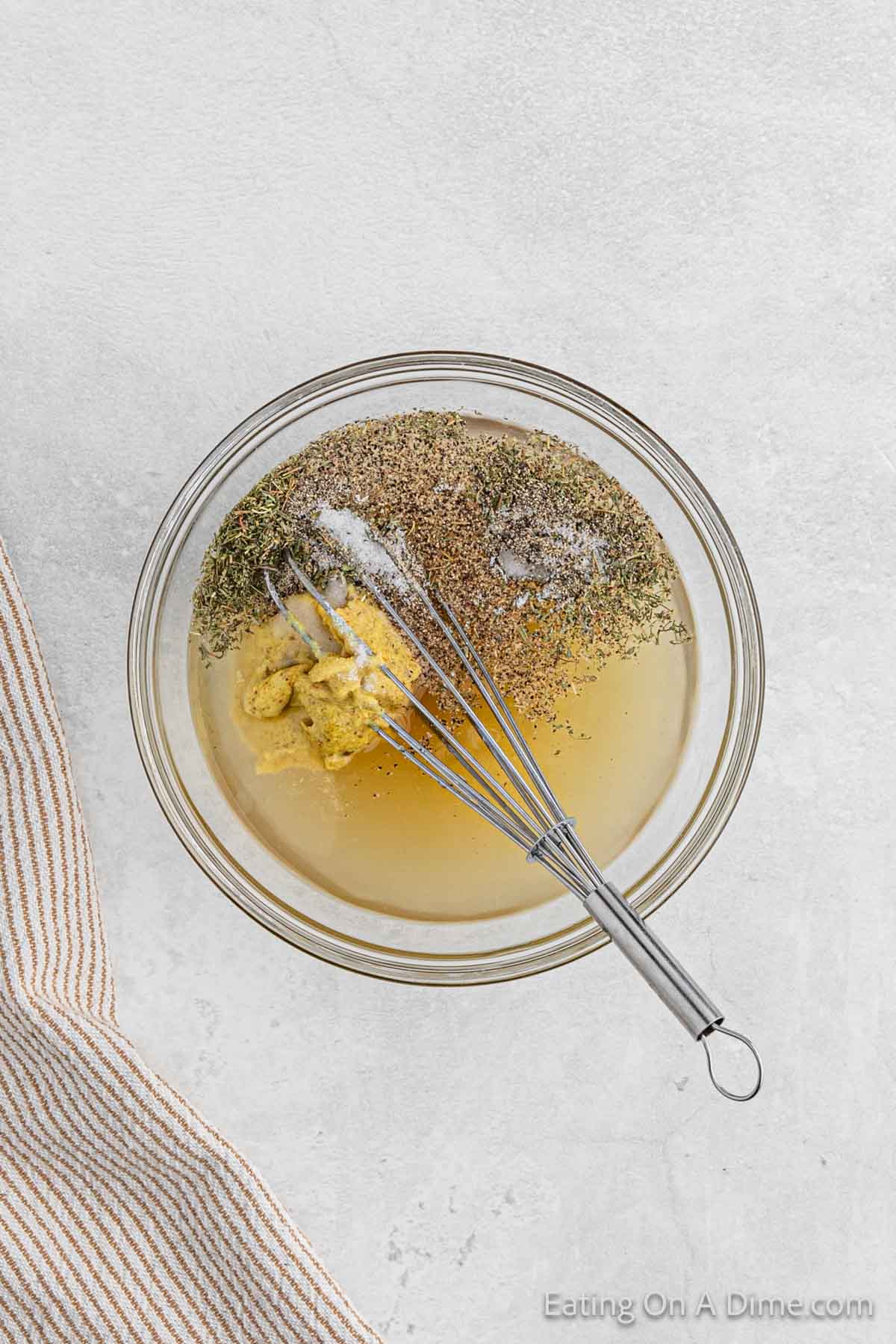 A clear glass bowl contains a mixture of olive oil, minced garlic, Dijon mustard, herbs, and spices, with a metal whisk resting inside the bowl. The bowl is on a light-colored surface next to a beige and white striped kitchen towel—perfect for prepping your Crock Pot dishes like kielbasa and potatoes.