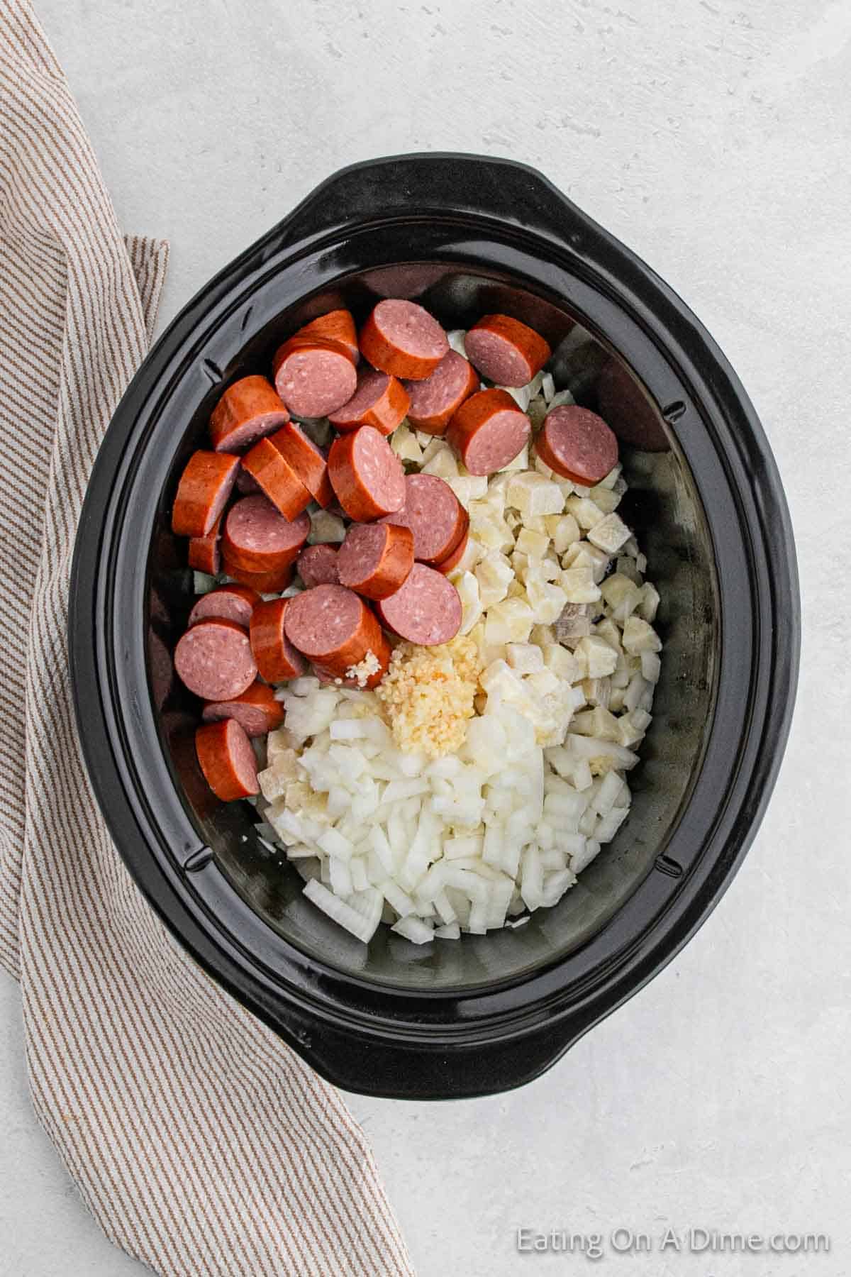 A slow cooker filled with sliced sausages, chopped onions, and minced garlic. Tender chunks of potatoes meld perfectly with the savory flavors. A beige and white striped cloth napkin is placed next to the Crock Pot on a light-colored surface.