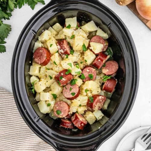 A Crock Pot filled with a hearty dish of cubed potatoes and slices of kielbasa, garnished with chopped parsley. The ingredients appear to be seasoned and combined, creating a savory meal. The cooker sits on a light-colored countertop with some herbs nearby.