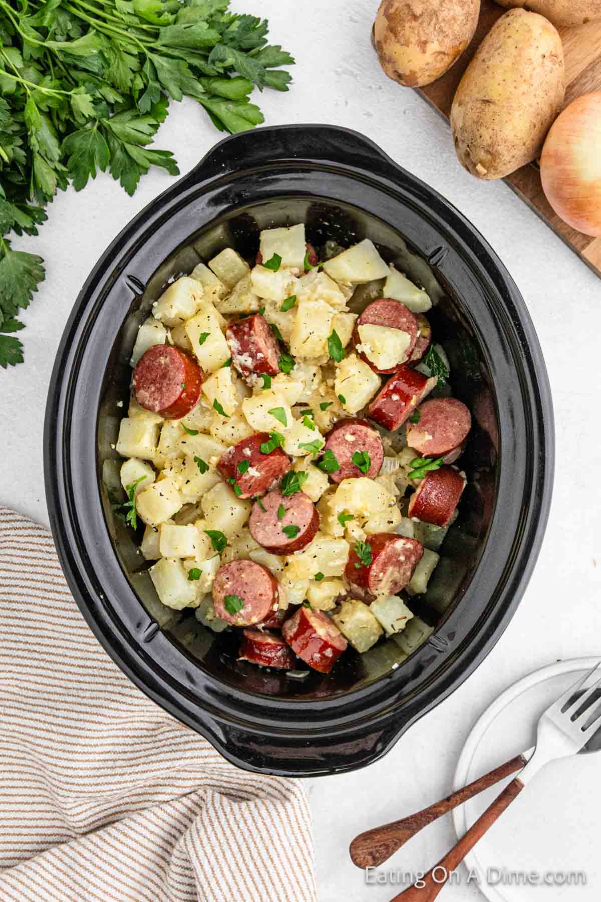 A Crock Pot filled with a hearty meal of sliced kielbasa, diced potatoes, and chopped onions, garnished with fresh parsley. Surrounding the pot are fresh parsley, whole potatoes, an onion, a striped cloth, wooden utensils, and a knife.