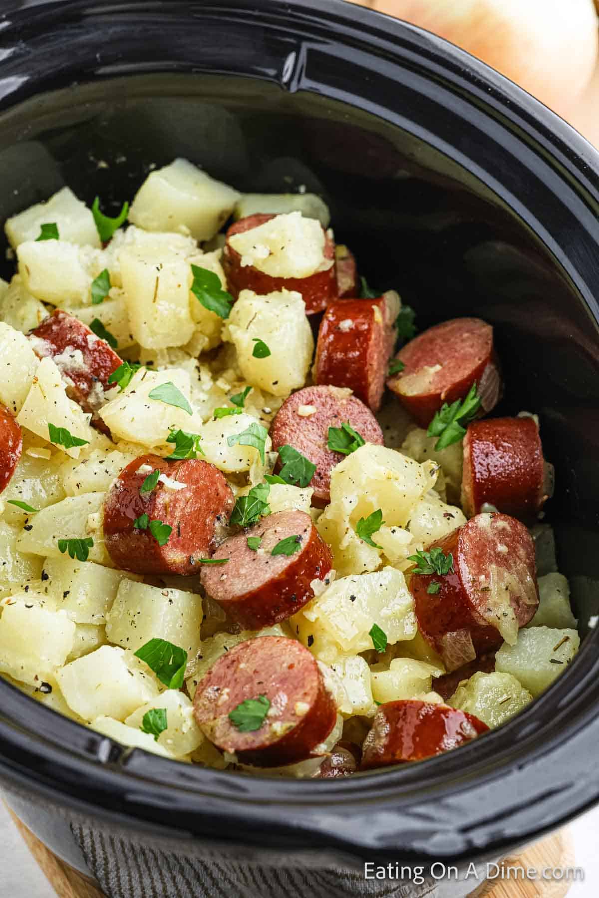 A close-up of a Crock Pot filled with a dish of diced potatoes and sliced kielbasa, garnished with chopped parsley. The mixture appears well-seasoned and cooked, presenting a hearty, rustic appearance.