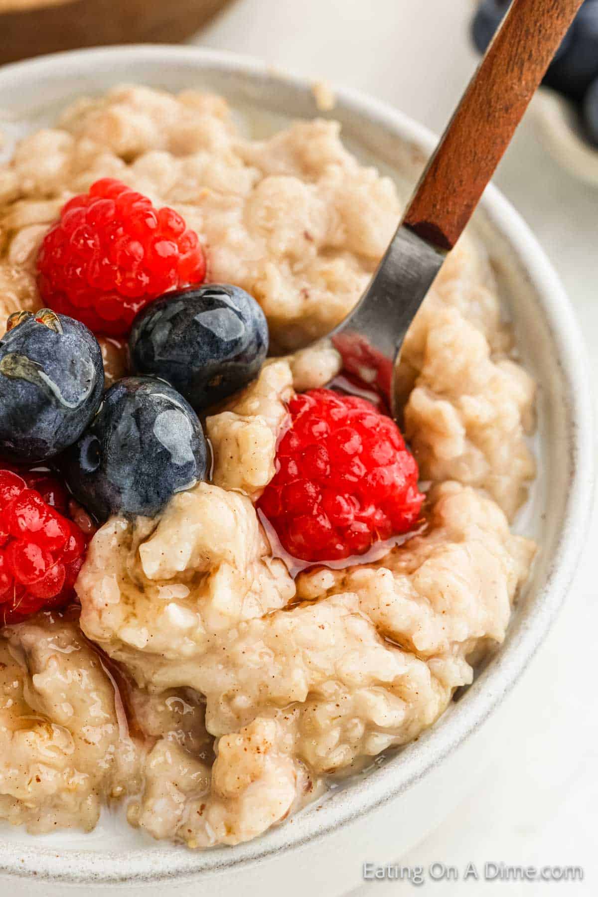 A close-up of a bowl of oatmeal topped with fresh raspberries and blueberries. A spoon is partially inserted into the oatmeal, ready for a bite. The background is slightly blurred, emphasizing the texture and colors of the crock pot-cooked oatmeal and vibrant berries.