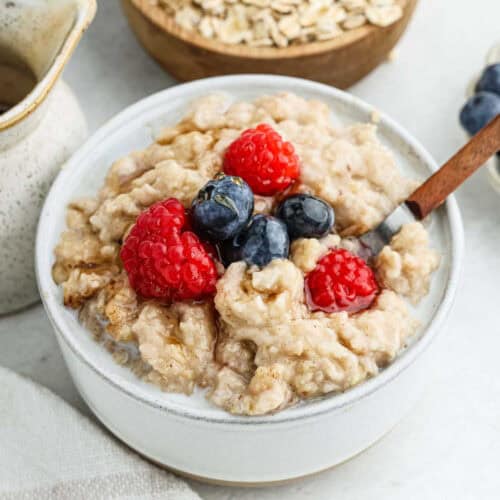 A bowl of creamy oatmeal, prepared effortlessly in a crock pot, is topped with fresh raspberries and blueberries. A spoon sits in the bowl, and a small jug of milk is visible to the side. A nearby bowl contains oats, and the background shows another small bowl with more blueberries.