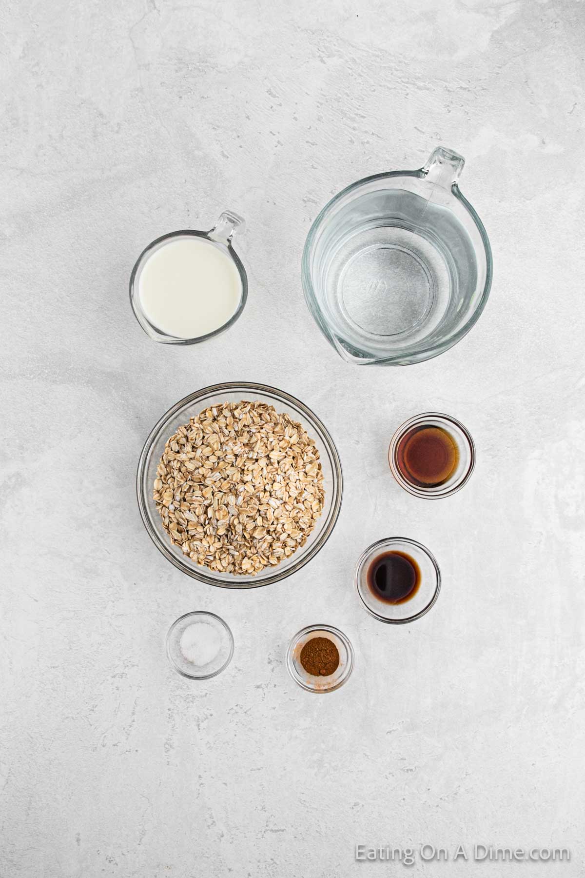 A top-down view of various ingredients in separate bowls and containers on a light gray surface. Ingredients for Crock Pot oatmeal include rolled oats in a glass bowl, a cup of milk, a cup of water, small bowls containing vanilla extract, and cinnamon.