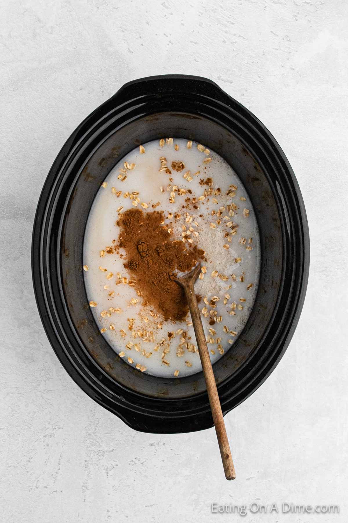 A crock pot with a mixture of oats, milk, and a heap of cinnamon in the center. A wooden spoon is placed inside the pot. The crockpot sits on a light-colored surface, ready to serve up warm oatmeal goodness.